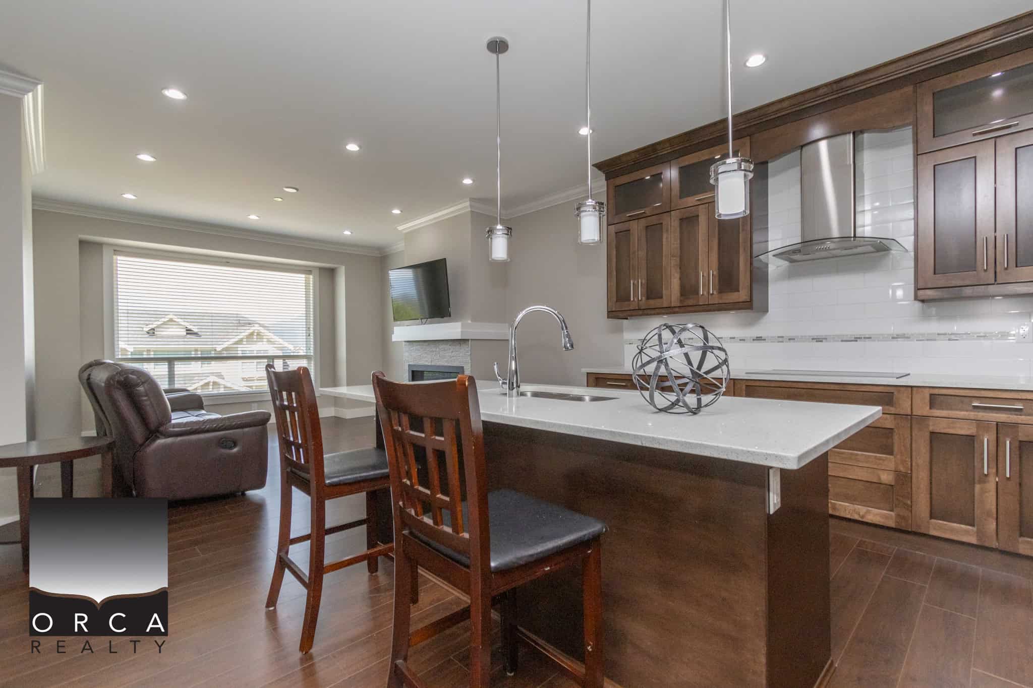 Bright modern kitchen with dark hardwood cabinets, stainless steel appliances, and a spacious white countertop island in a contemporary home. Large window lets in natural light, with cozy living area and fireplace nearby.