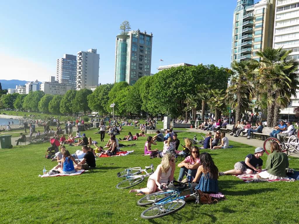 Vibrant urban park with people relaxing on grass, cycling, and enjoying sunny weather against a city skyline with high-rise buildings and lush green trees.