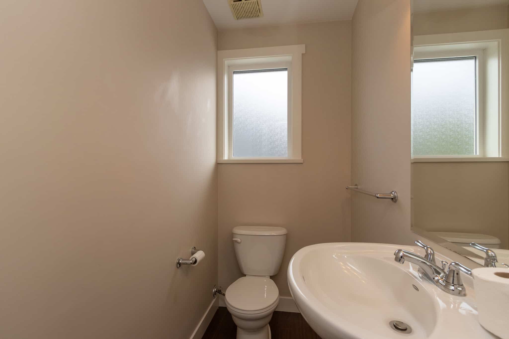 IMG_1783 Bright beige bathroom interior with a white toilet, large mirror, and a white pedestal sink, featuring two frosted windows for natural light, modern fixtures, and clean minimalist design.