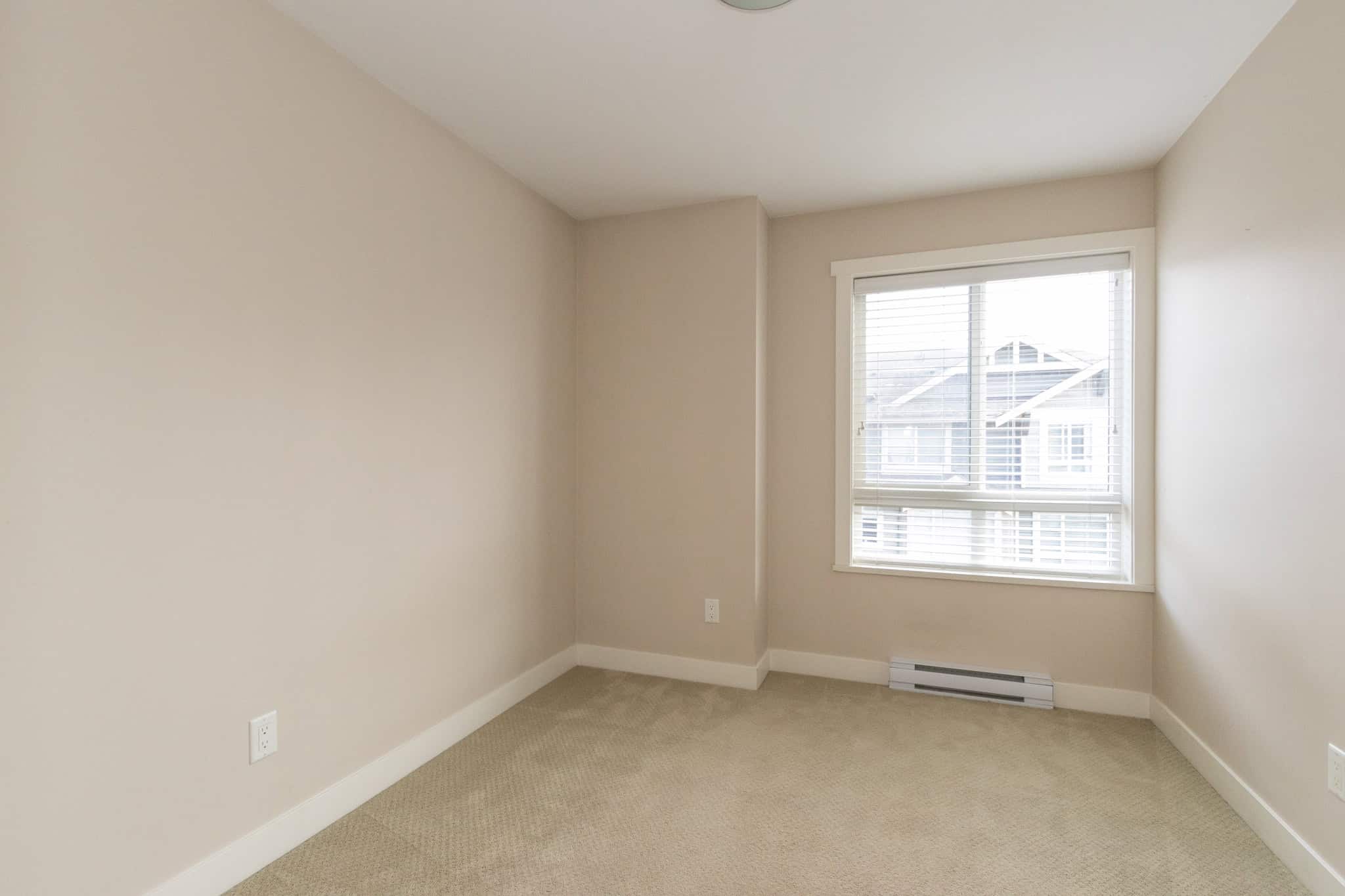 IMG_1820 Bright, empty bedroom with neutral walls and carpet, featuring large window with white blinds offering natural light, perfect for Vancouver area homebuyers interested in Orca Realty Inc. listings.
