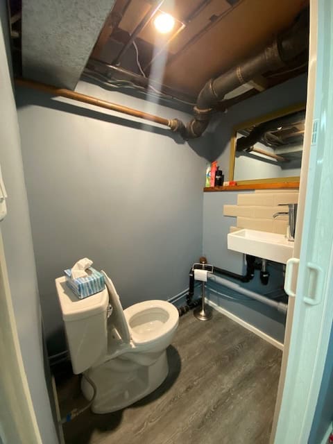 A bathroom with a toilet and small sink, exposed plumbing, grey walls, and industrial ductwork on the ceiling, ideal for small or basement spaces.