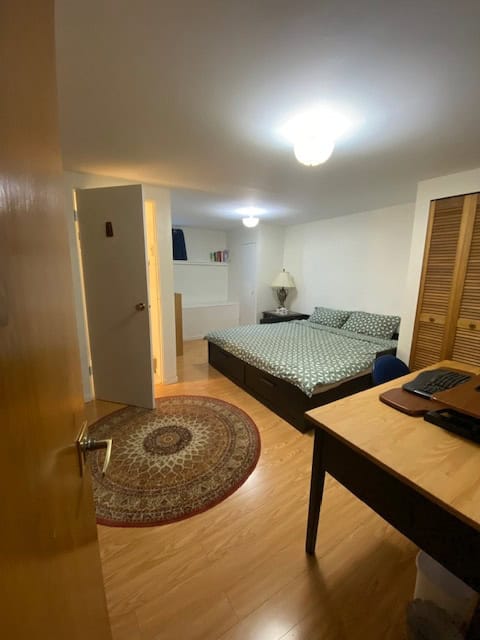 Modern furnished bedroom with a cozy bed, wooden flooring, a small rug, and a professional workspace - ideal for comfortable living and remote work in a Vancouver area home.