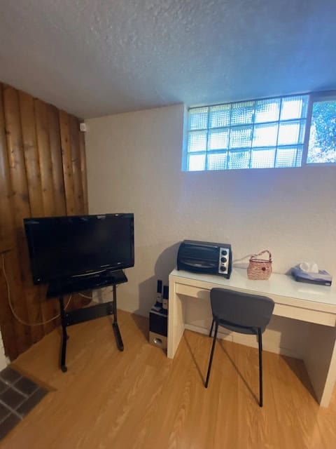 Cozy home office with a television on a stand, white desk with a black chair, toaster oven, and decorative wicker basket in a bright, sunlit room with a small window and wood-paneled wall.