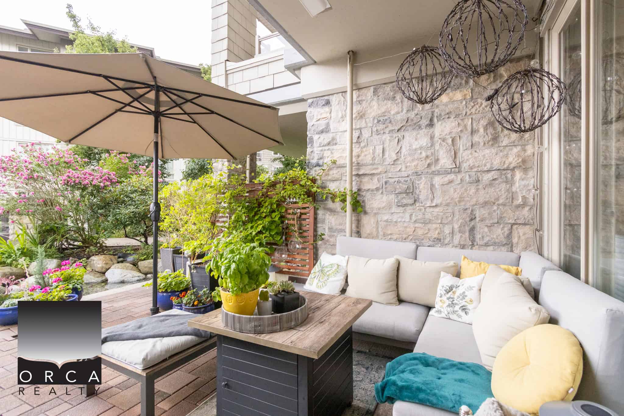 Cozy outdoor patio with comfortable sectional sofa, potted plants, decorative hanging lights, and a large umbrella on a sunny day, perfect for relaxing and entertaining at a Vancouver-area home.