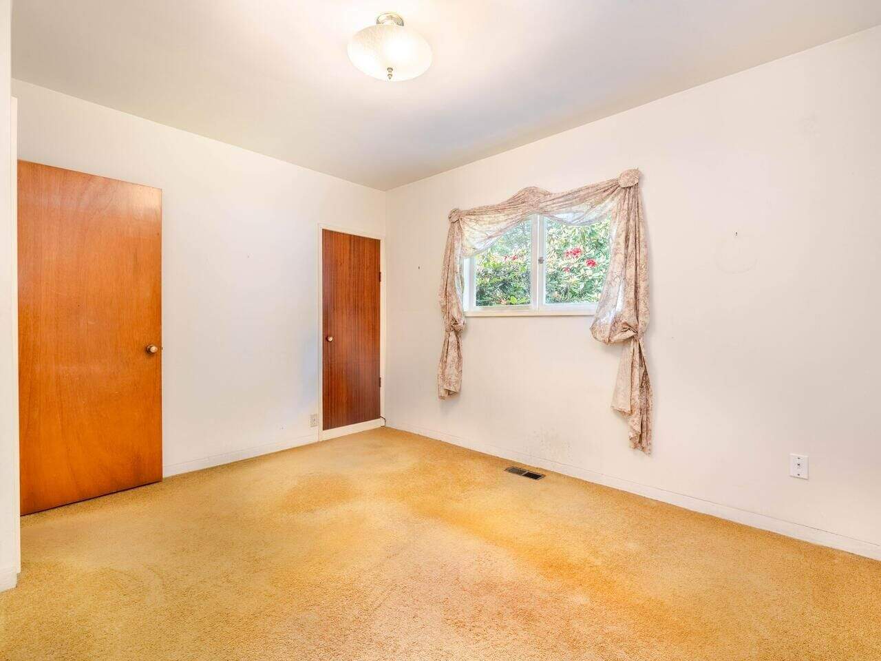 Bright empty bedroom with beige carpet, white walls, and a window with floral curtains in a home listed by Orca Realty Inc. in Victoria, BC. Perfect for homebuyers seeking a cozy, affordable property.