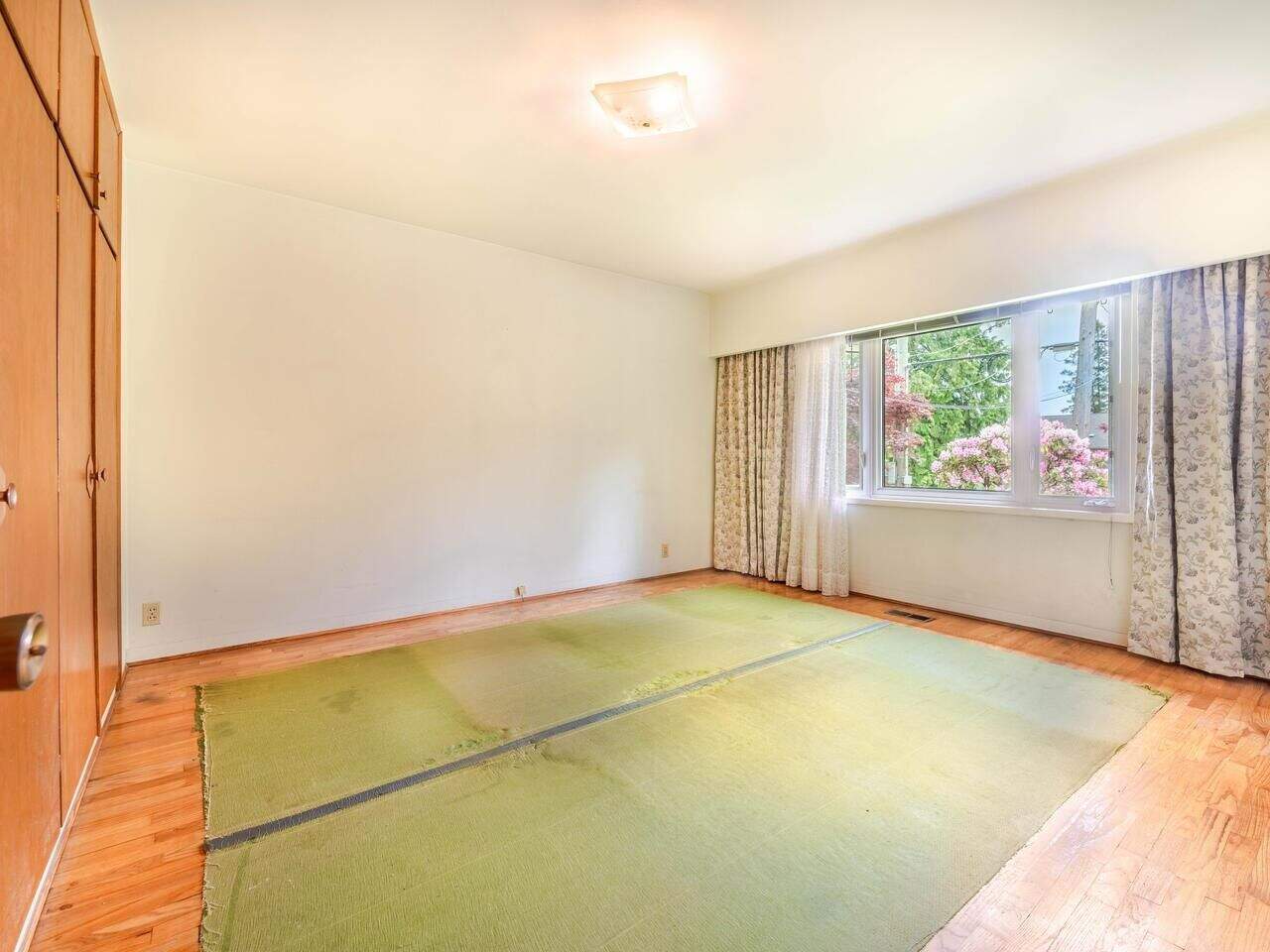 Floor-to-ceiling window in a bright and spacious bedroom with wooden floorboards, vintage curtains, and a green rug, showcasing a cozy and inviting living environment ideal for resale or rental properties in Victoria, BC.