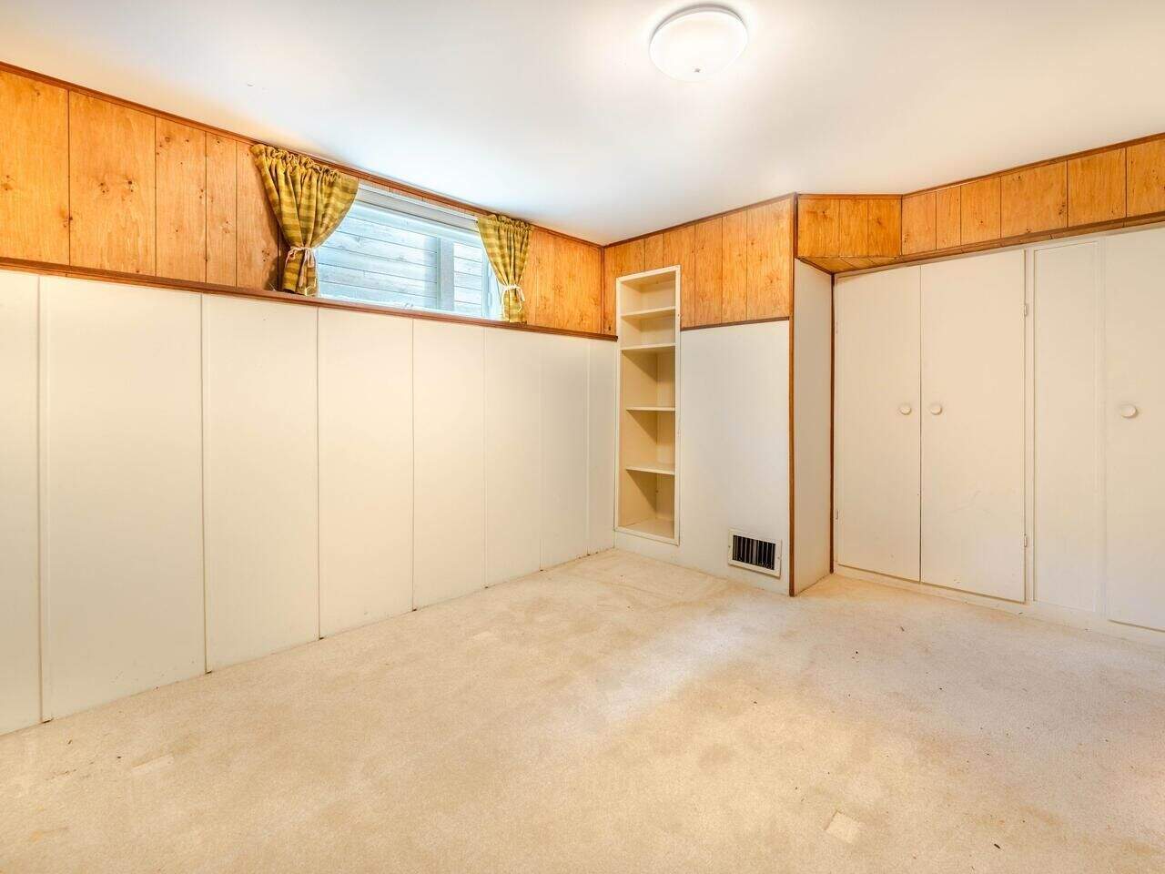 Spacious basement bedroom with wood-paneled walls, built-in shelves, and a small window with yellow plaid curtains, perfect for a cozy home in Vancouver.