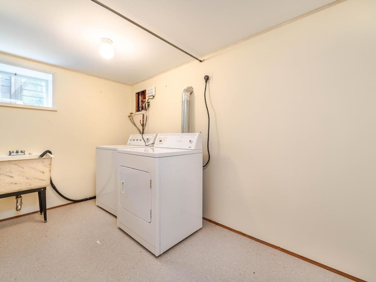 Large laundry room with washer and utility sink in a residential basement, featuring neutral walls, small basement window, and simple lighting. Ideal for home laundry and storage solutions.