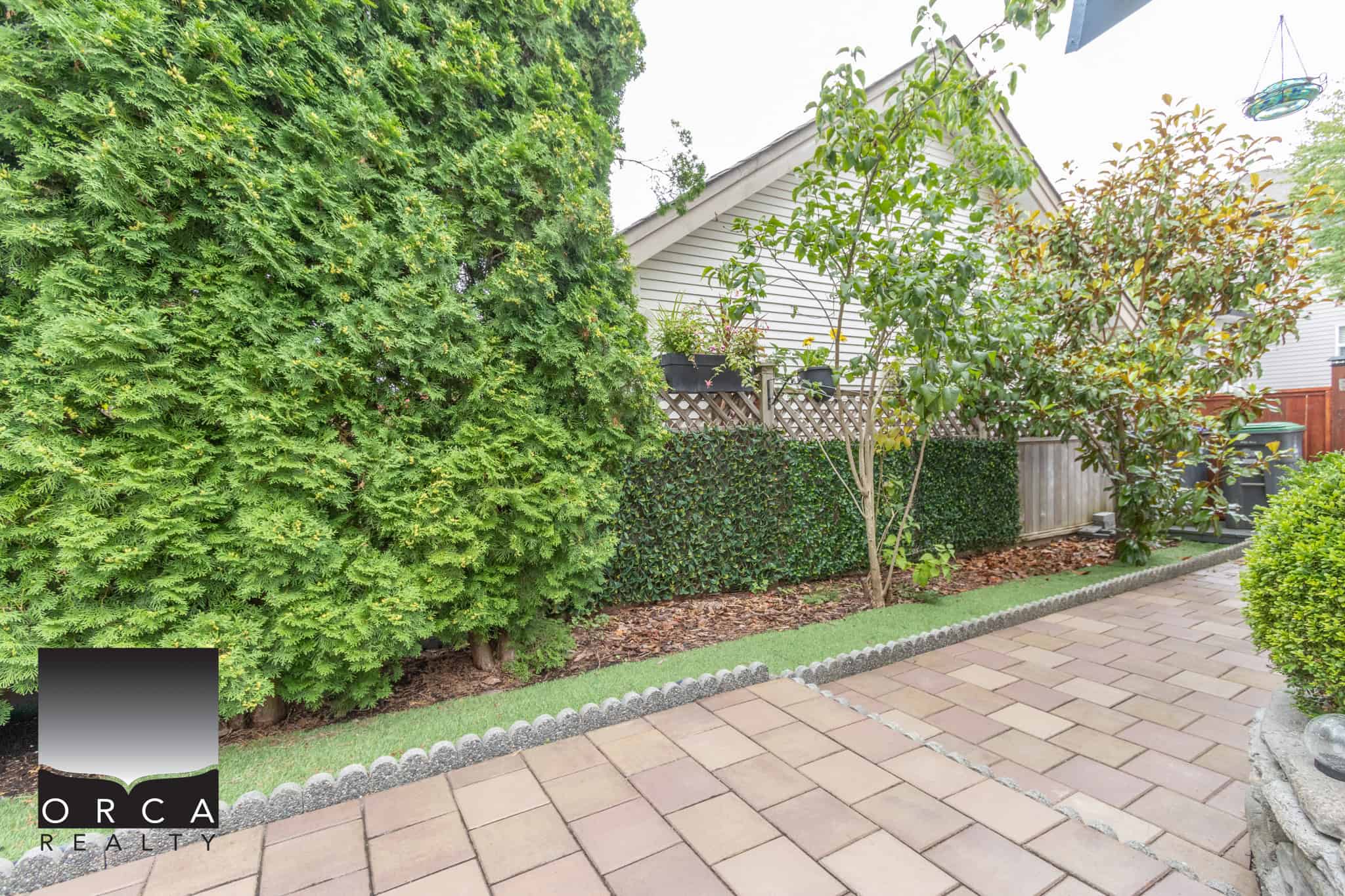 Lush green backyard garden with well-manicured shrubs, trees, and a paved patio area, ideal for outdoor living and relaxation, featured on Orca Realty Inc. website.