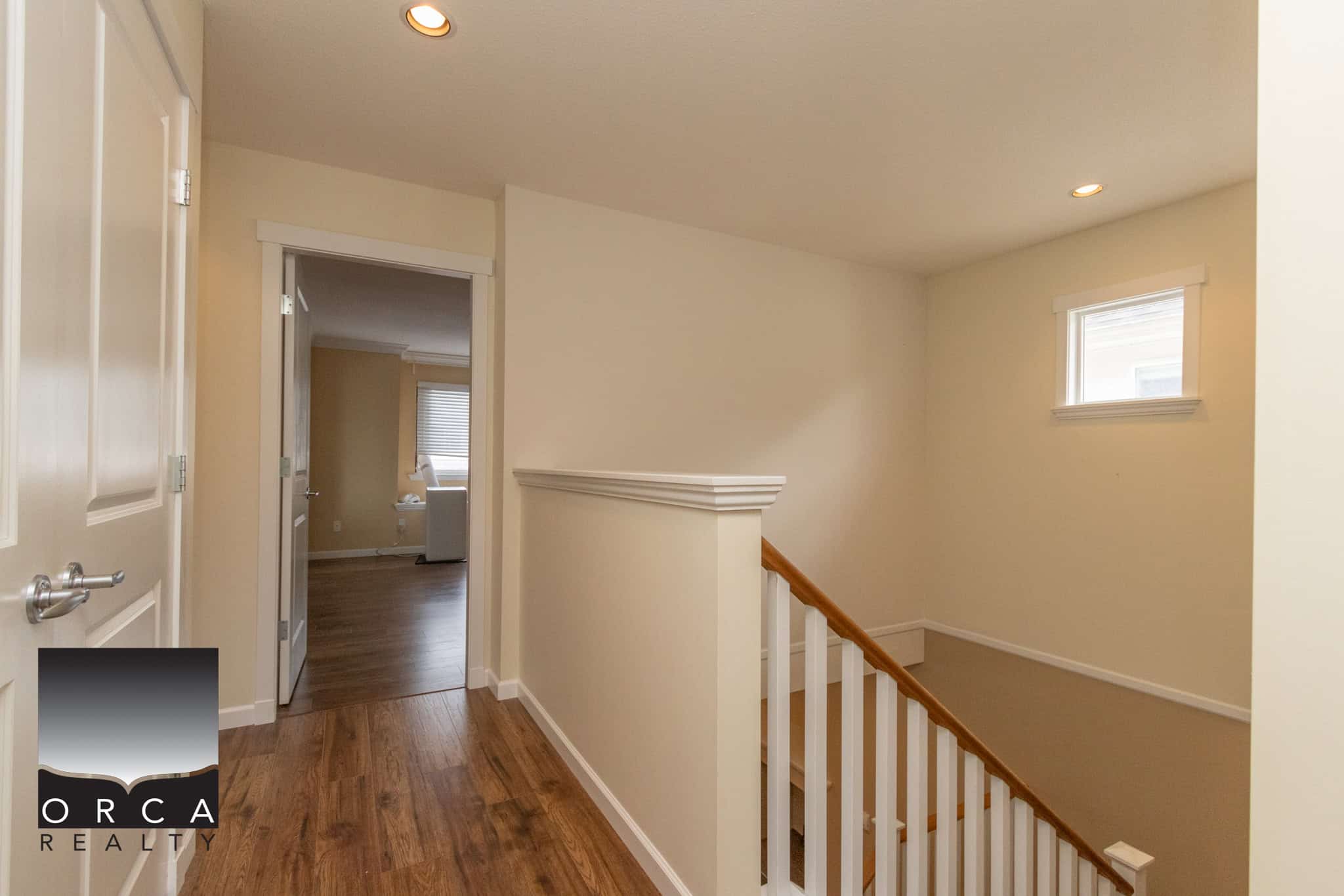 Bright, modern upstairs hallway with hardwood flooring, natural light from small window, and white walls, showcasing a cozy residential interior by Orca Realty Inc.