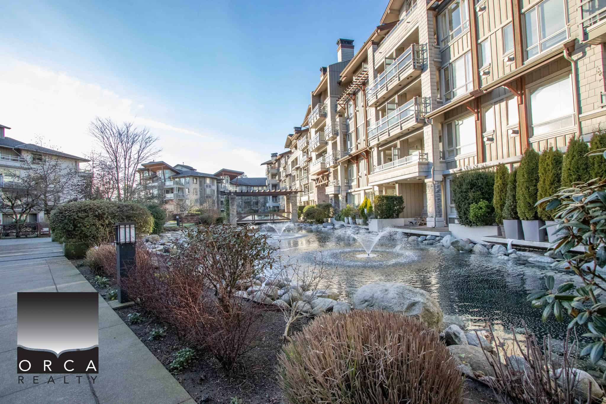 Serene residential courtyard with modern apartment buildings, landscaped gardens, water features, and outdoor seating in an upscale community managed by Orca Realty Inc.