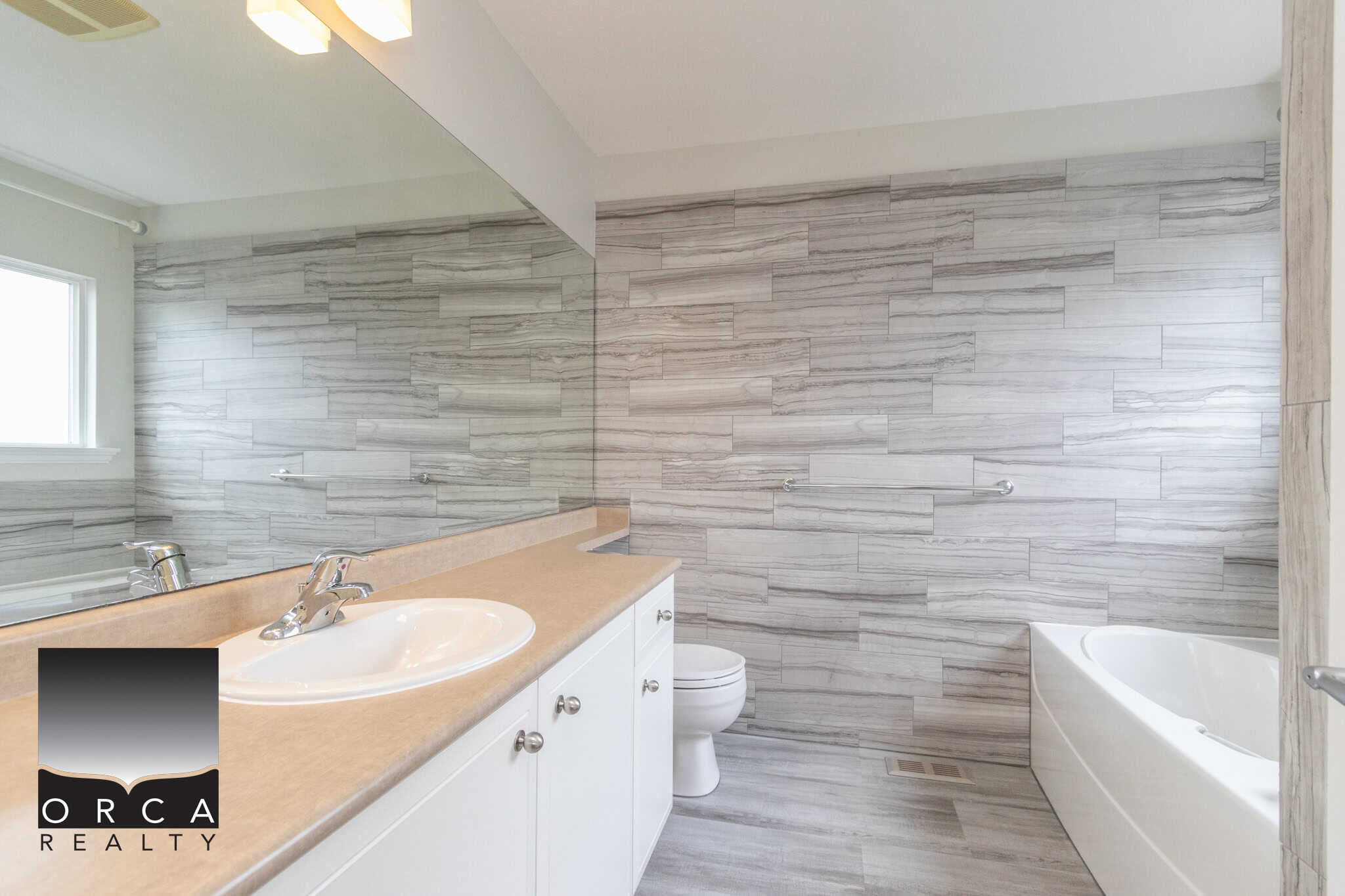 Modern bathroom with grey stone tile wall, white vanity with beige countertop, bathtub, and window, showcasing upscale home design by Orca Realty Inc. for Vancouver real estate.