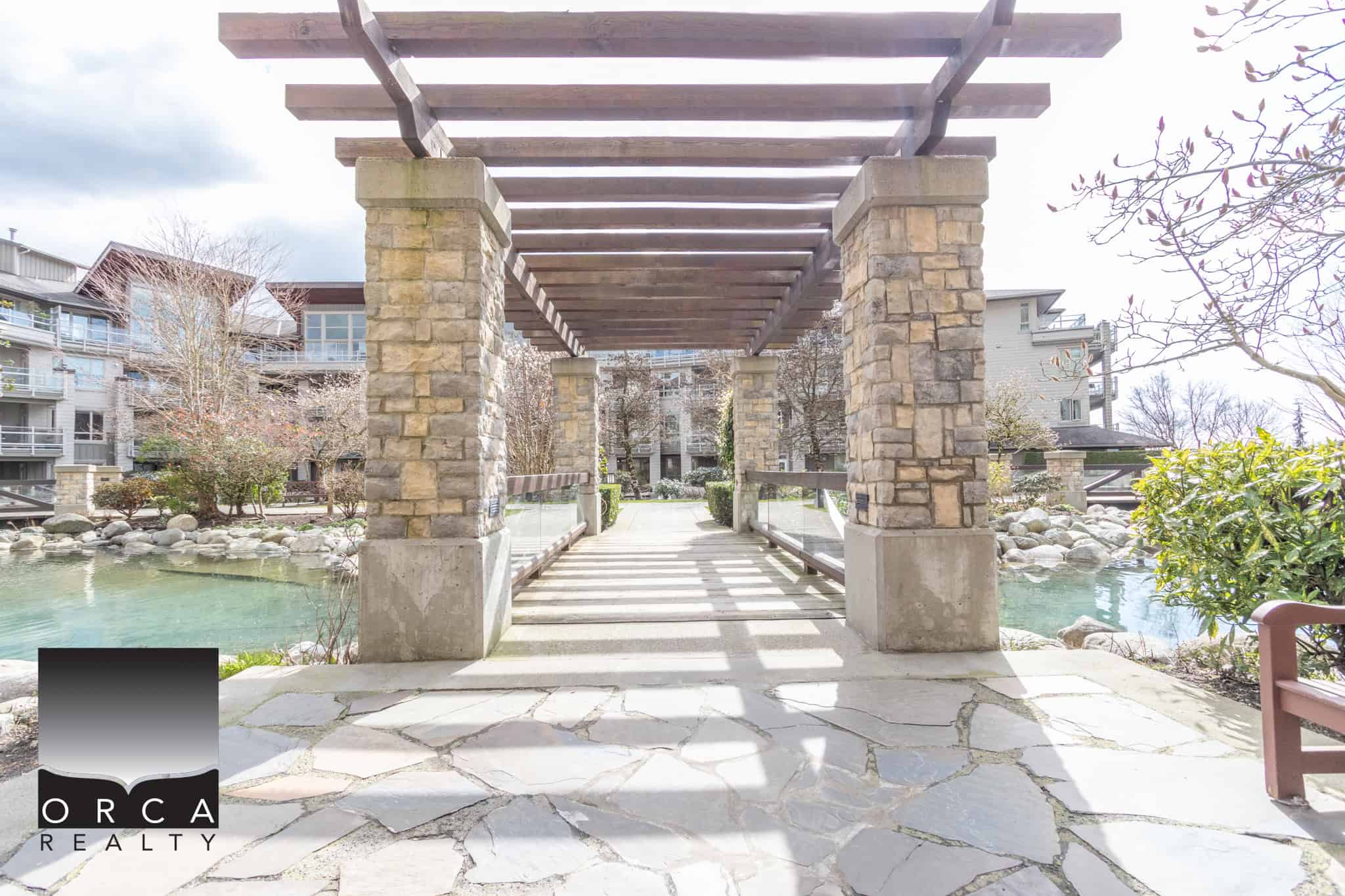 Stone arch bridge over pond at Orca Realty Inc. residential community in Vancouver, BC. Modern landscaping and serene water features create a peaceful environment for residents and visitors.