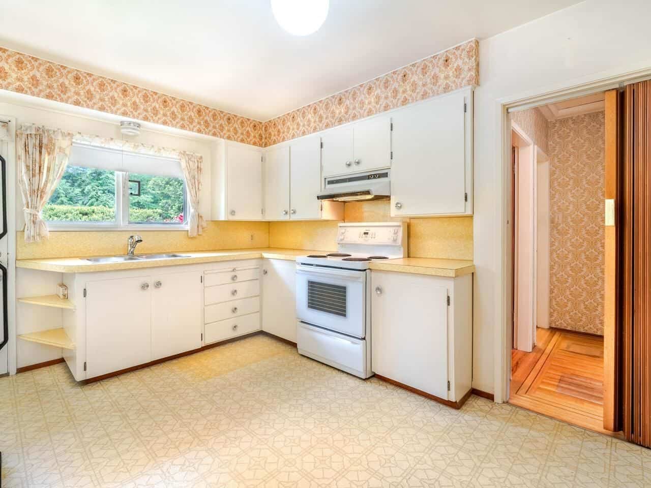 White kitchen with vintage yellow countertops and patterned wallpaper, featuring a window overlooking greenery, ideal for Vancouver homebuyers, real estate, and kitchen renovation keywords.