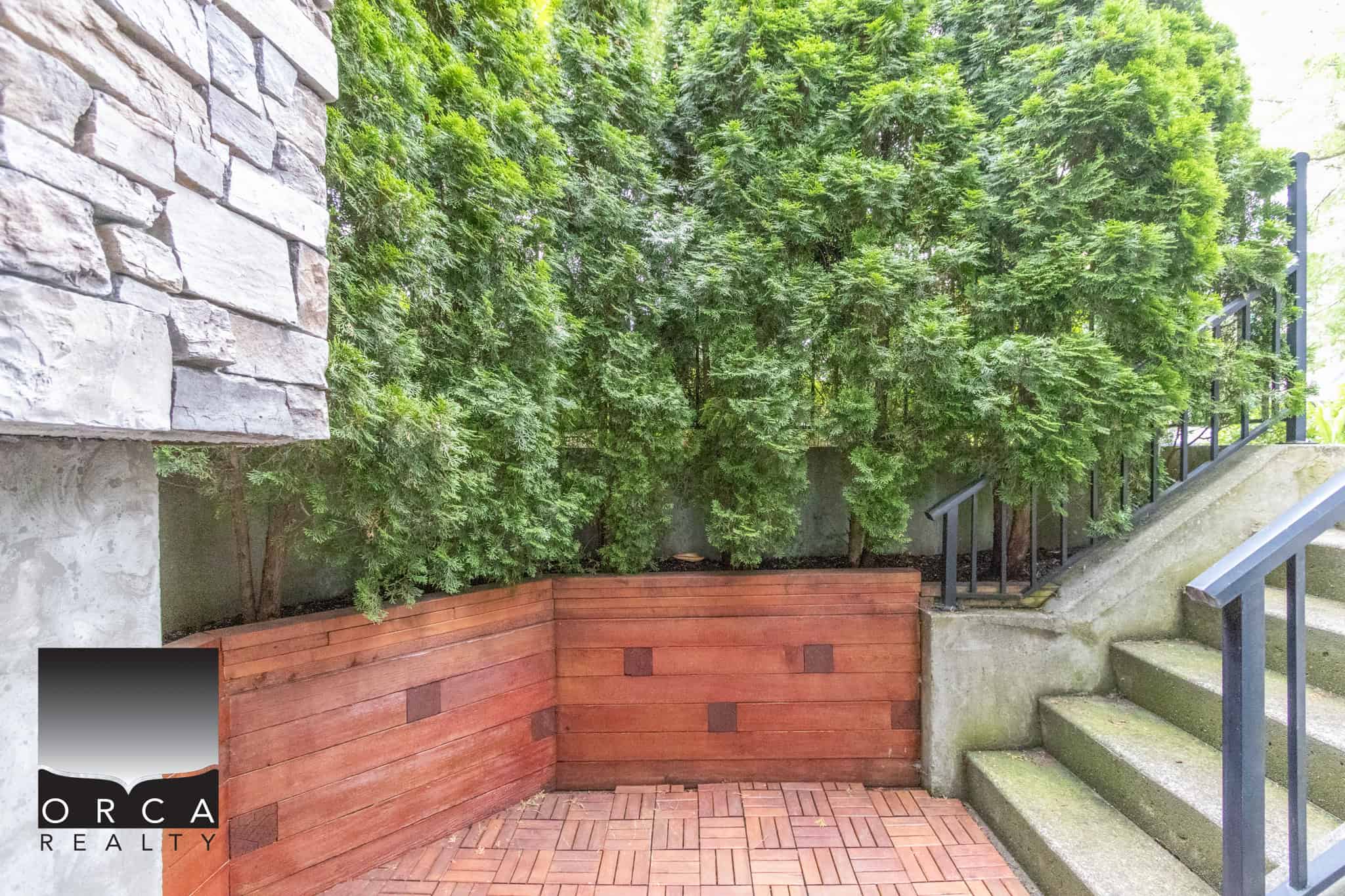 Lush green privacy hedge with concrete stairs and red brick planter at the entrance of a home, showcasing landscaping and outdoor design by Orca Realty Inc.