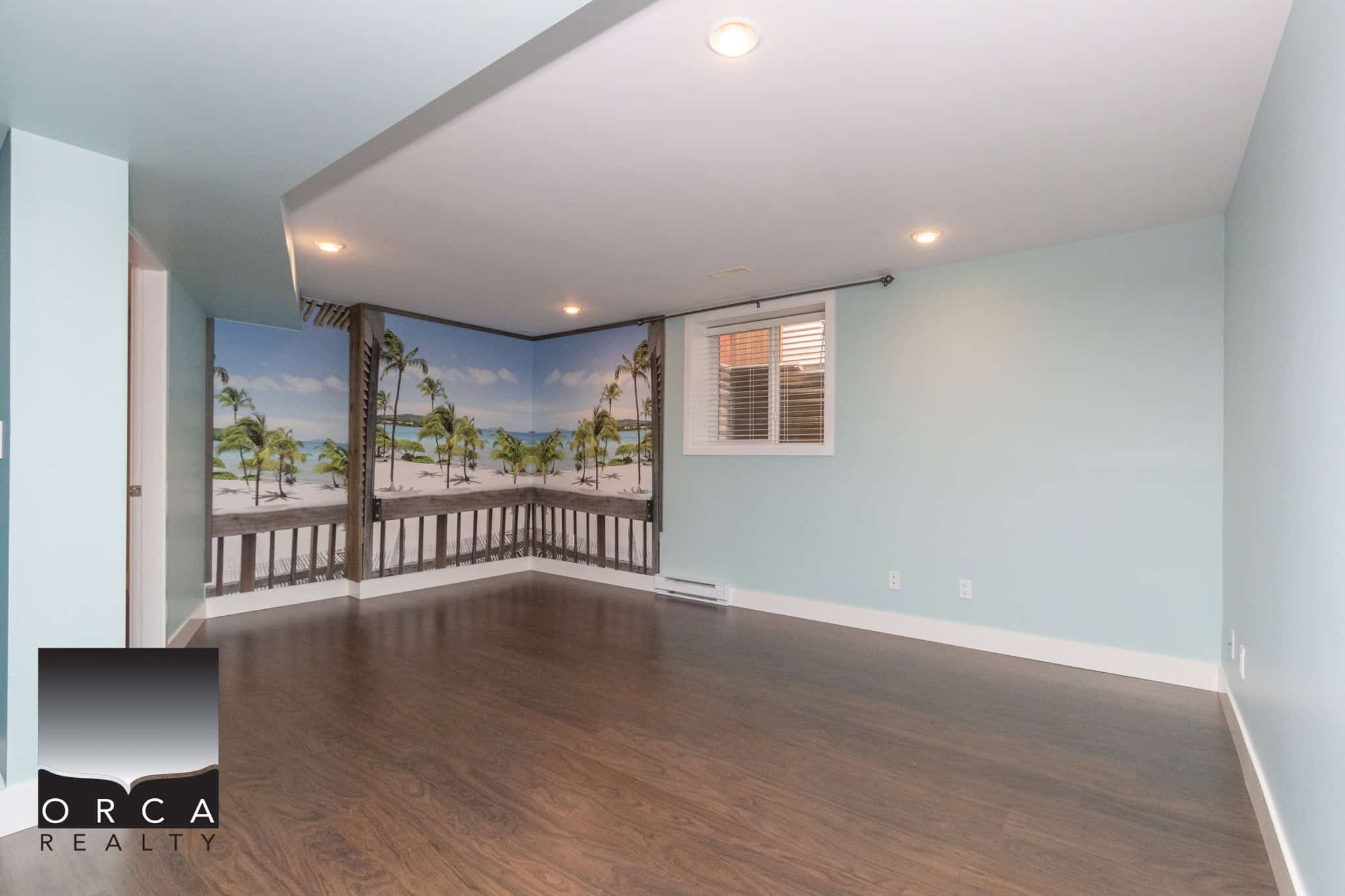 Beach-themed accent wall in a bright, spacious living room with hardwood flooring, large window, and recessed lighting, ideal for real estate listing photos.