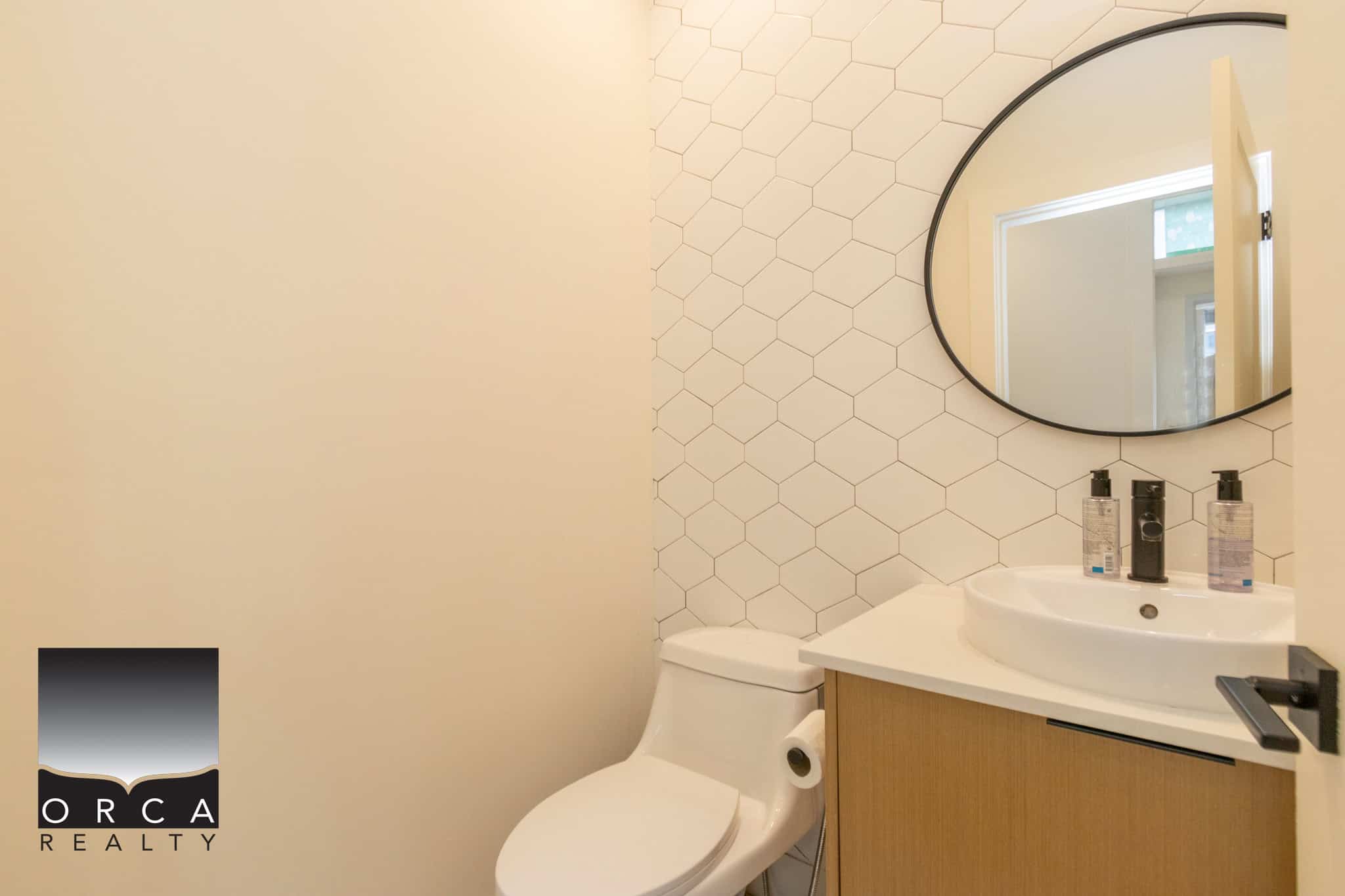 Modern bathroom with a white toilet, wood vanity, hexagon tile accent wall, and circular mirror, showcasing contemporary home design and amenities by Orca Realty Inc.