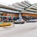 Modern mixed-use building with retail and residential units, featuring outdoor seating with umbrellas, parking lot, and contemporary design, located in a vibrant urban neighbourhood.