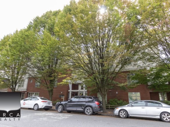 Lush green trees shade a modern residential building with red brick and white siding, with luxury cars parked along the street, showcasing Vancouver real estate and neighbourhood charm.