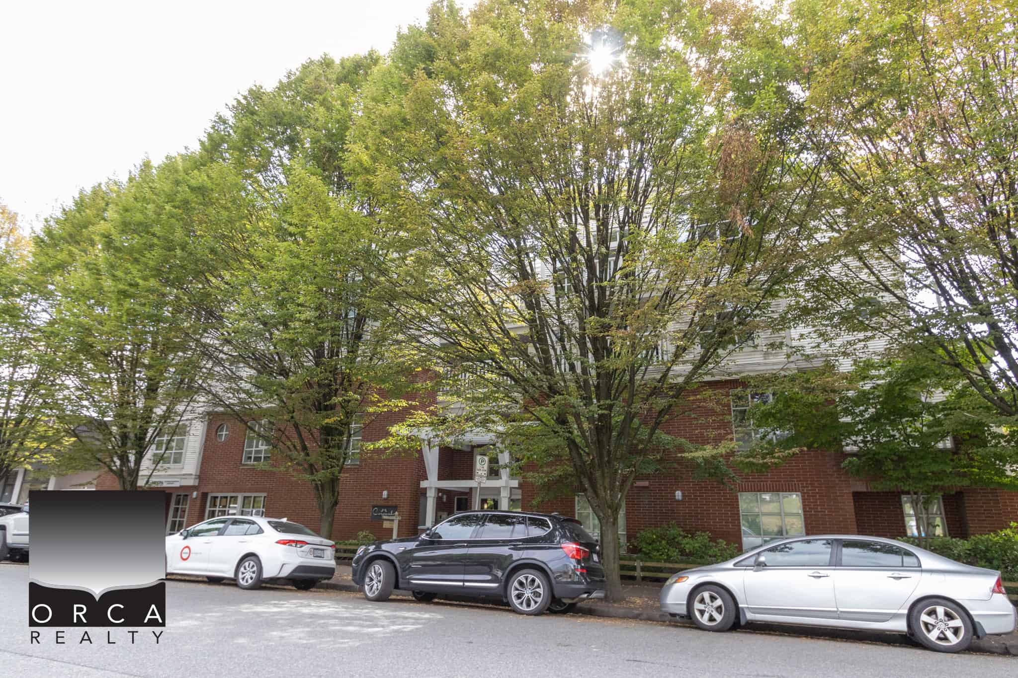 Lush green trees shade a modern residential building with red brick and white siding, with luxury cars parked along the street, showcasing Vancouver real estate and neighbourhood charm.