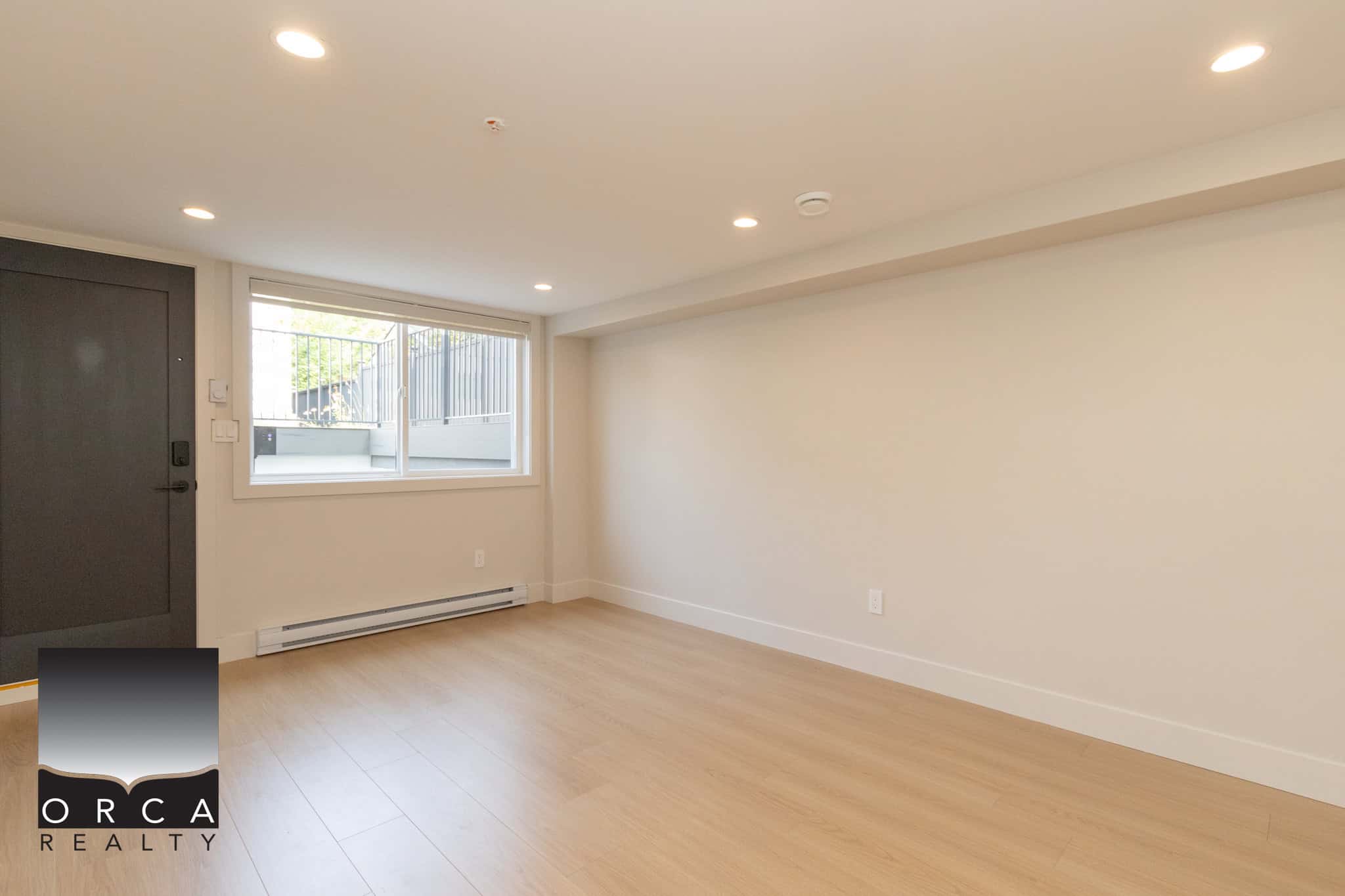 Bright, spacious interior of a modern apartment available through Orca Realty Inc., featuring light-toned hardwood flooring, large window with natural light, and minimalist design.