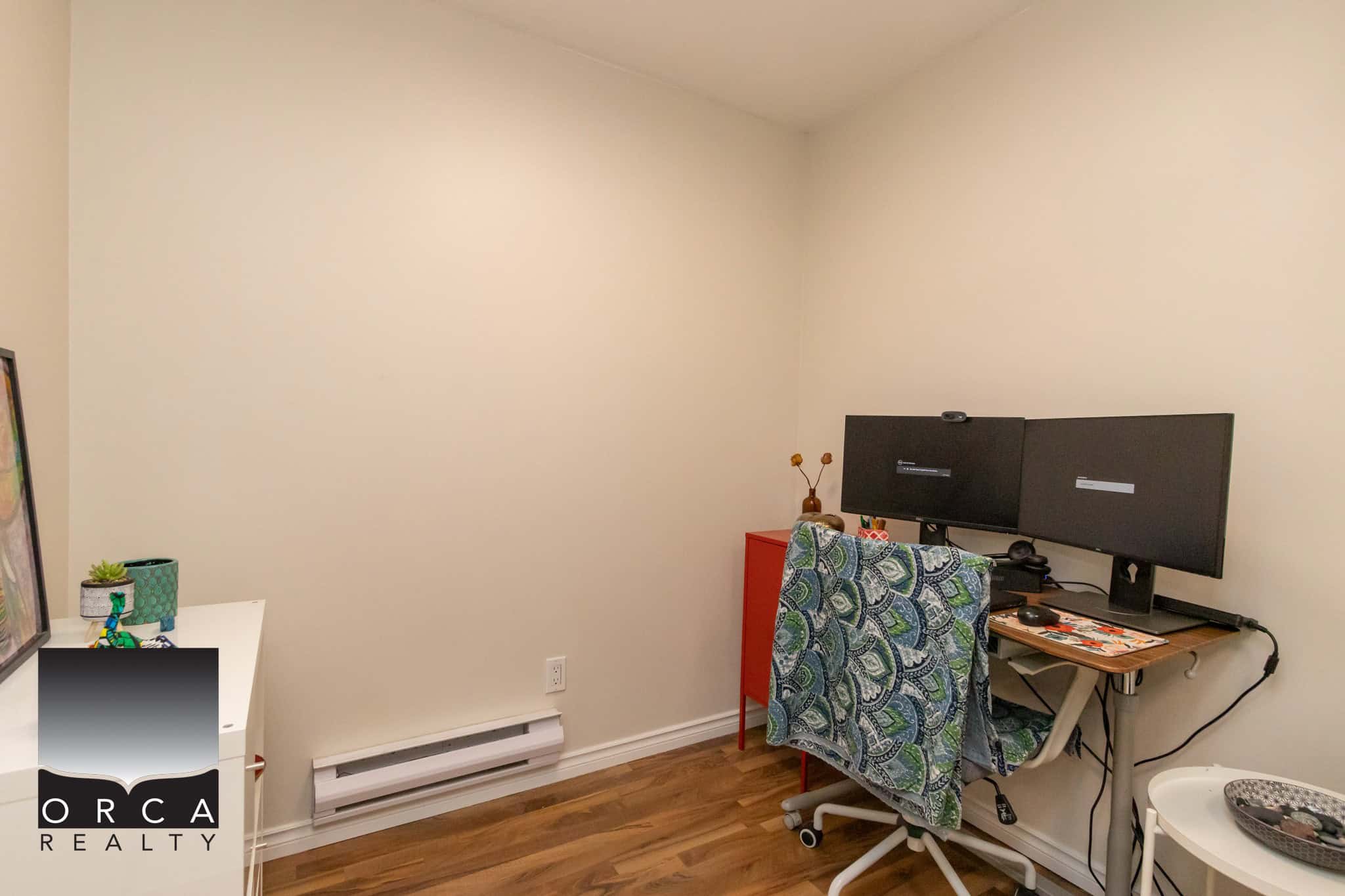 Modern home office with dual computer monitors, cozy decor, and hardwood flooring, ideal for remote work or study; part of a property listed by Orca Realty Inc. in Victoria, BC.