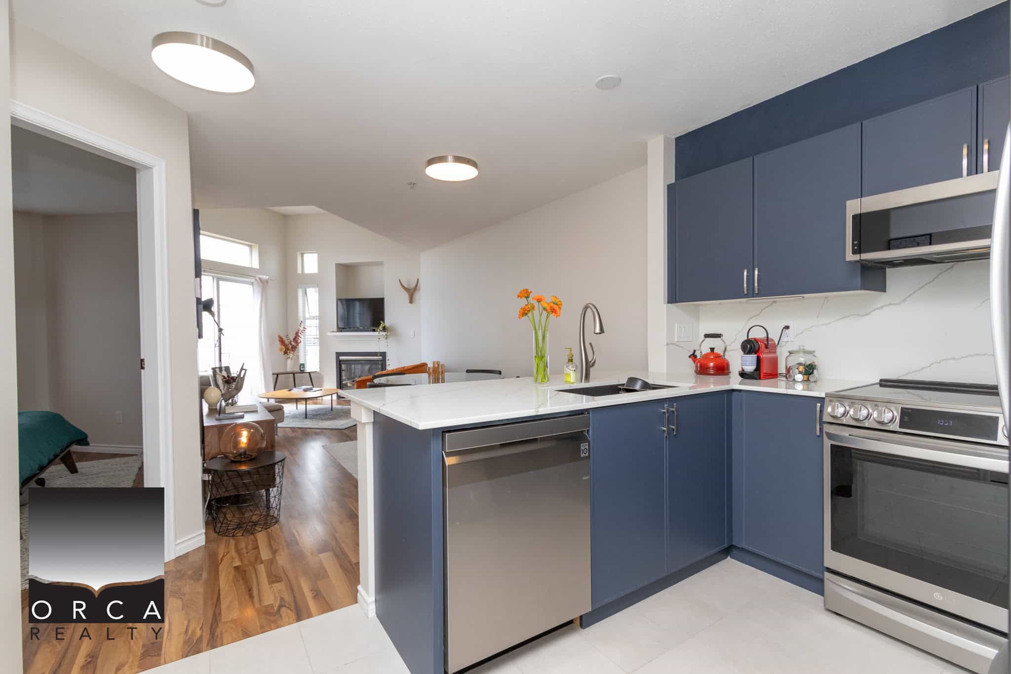 Contemporary blue kitchen with a white quartz countertop, stainless steel appliances, and modern cabinetry, overlooking a cozy living room with a fireplace and large windows.