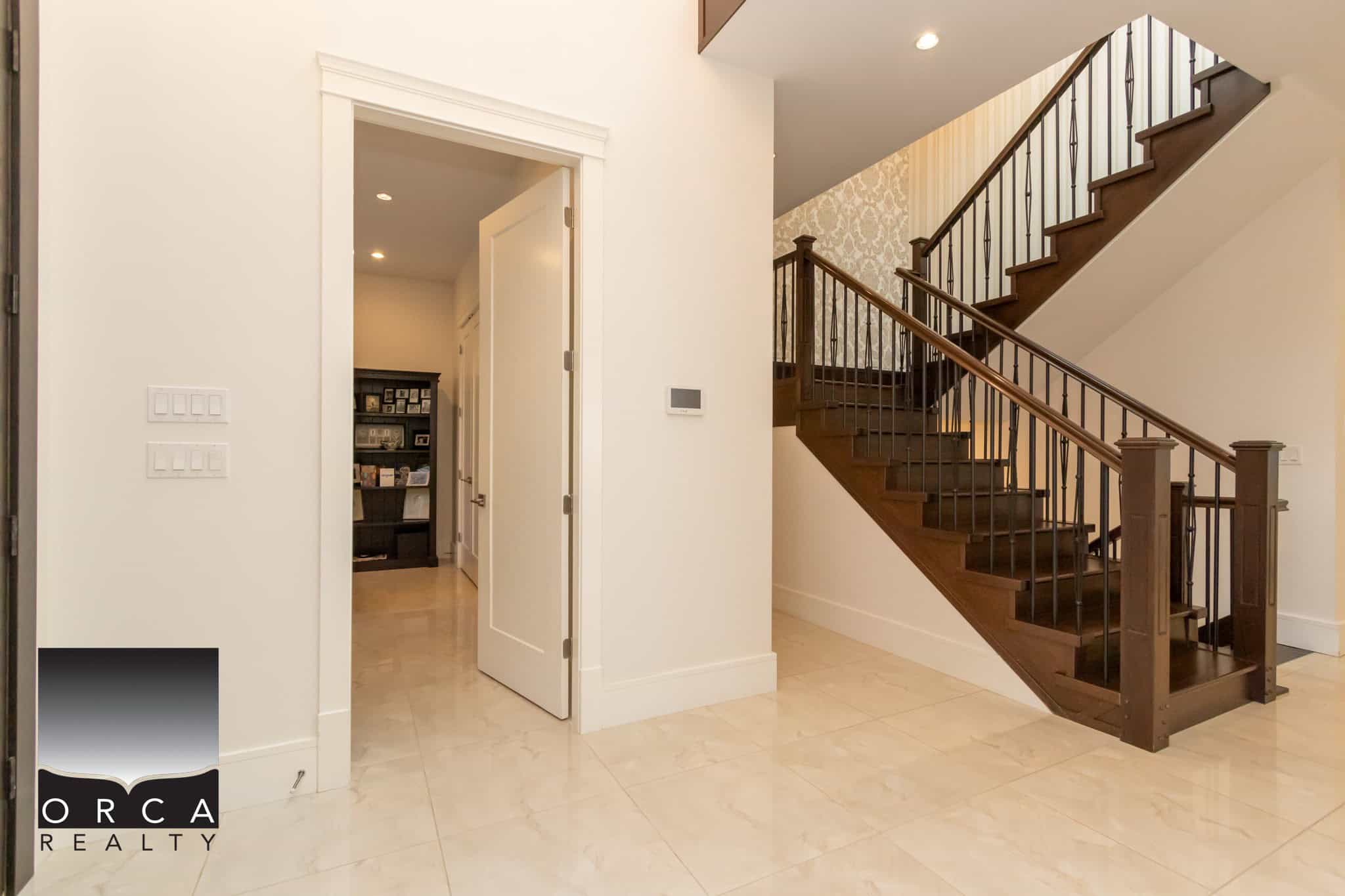 Spacious interior featuring a elegant staircase with wooden steps and black metal balusters, bright cream tiled flooring, and an inviting open doorway leading to a cozy home office or study area.