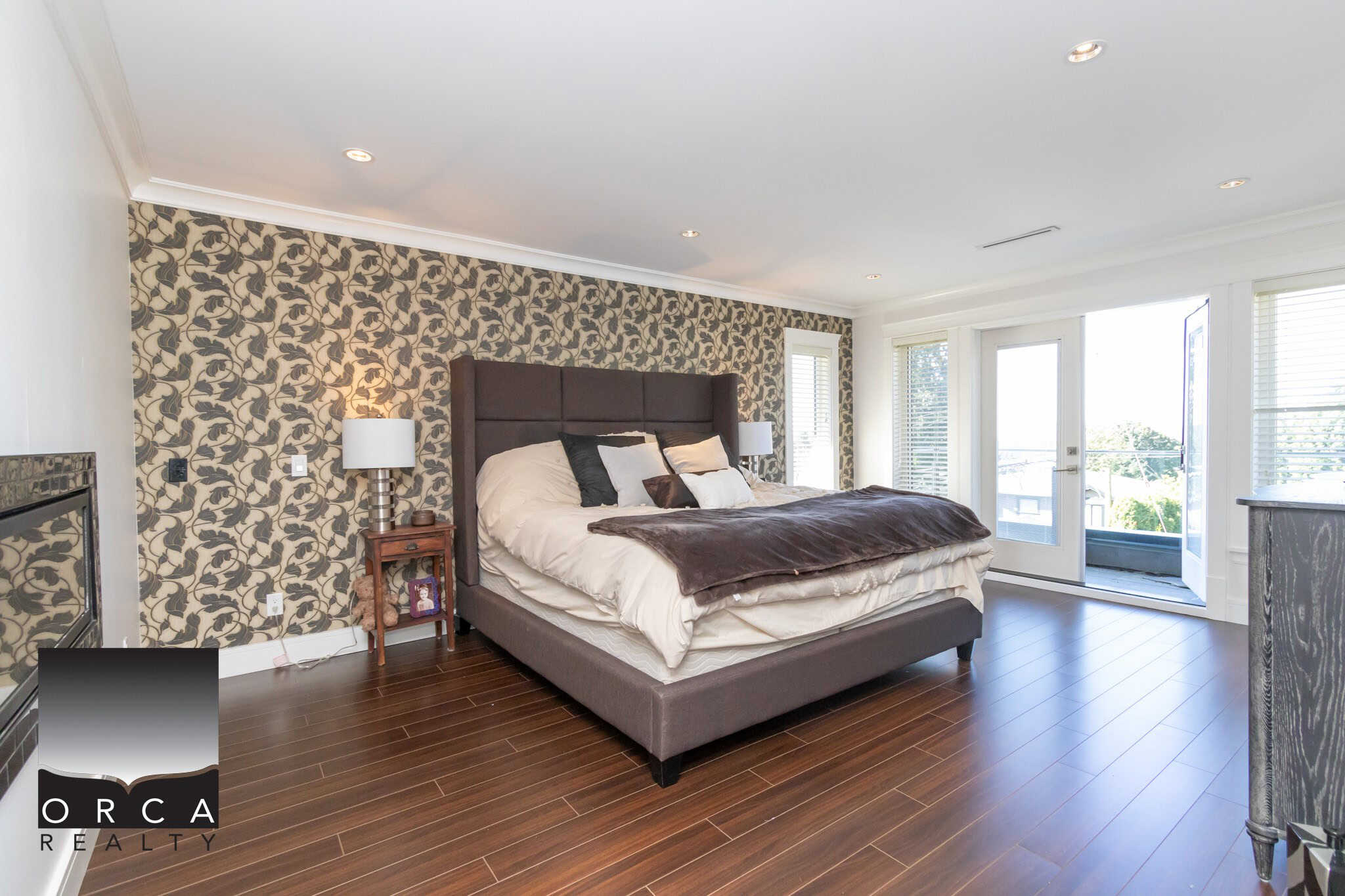 Modern bedroom with stylish wallpaper accent wall, large bed with plush bedding, natural light from balcony, hardwood flooring, perfect for Vancouver Island real estate.
