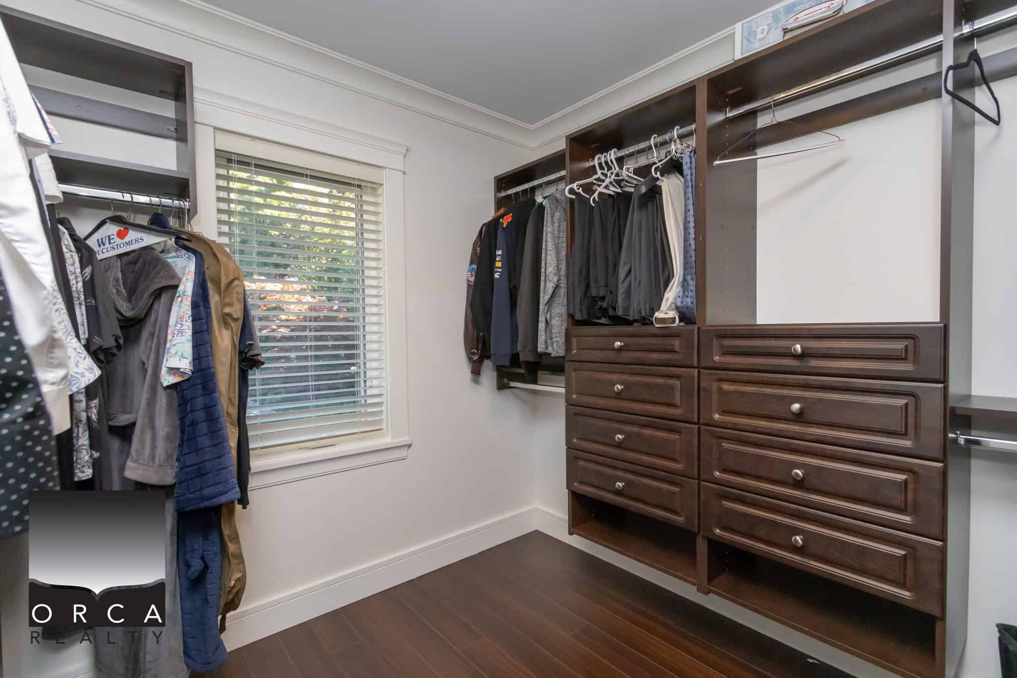 Spacious walk-in closet with organized clothing and wooden storage drawers, featuring white walls and a window bringing natural light into the space.