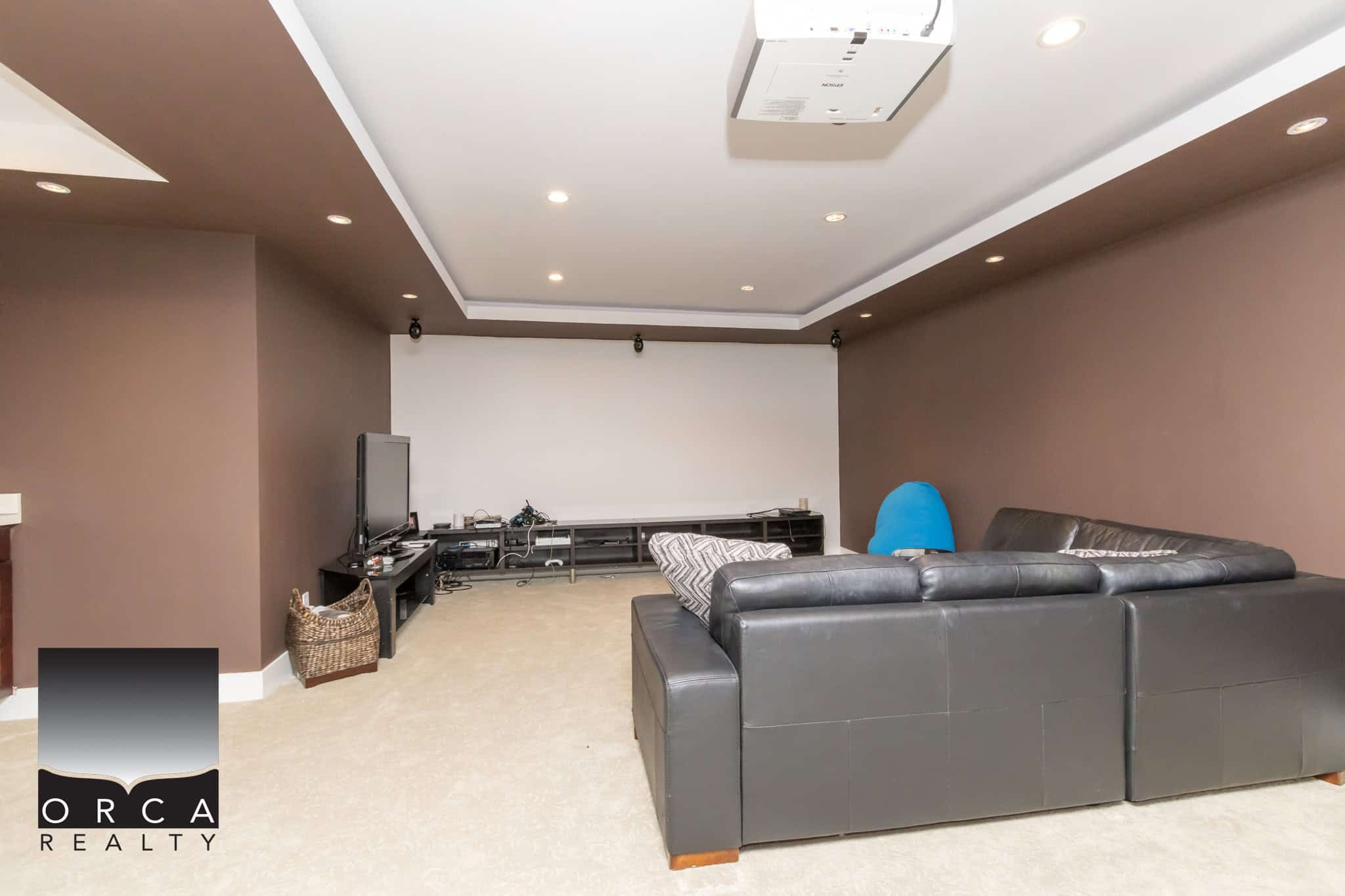 Spacious home theatre room with a large flat-screen TV, comfortable black leather sectional sofa, modern lighting, and audio-visual equipment, perfect for relaxing movie nights.