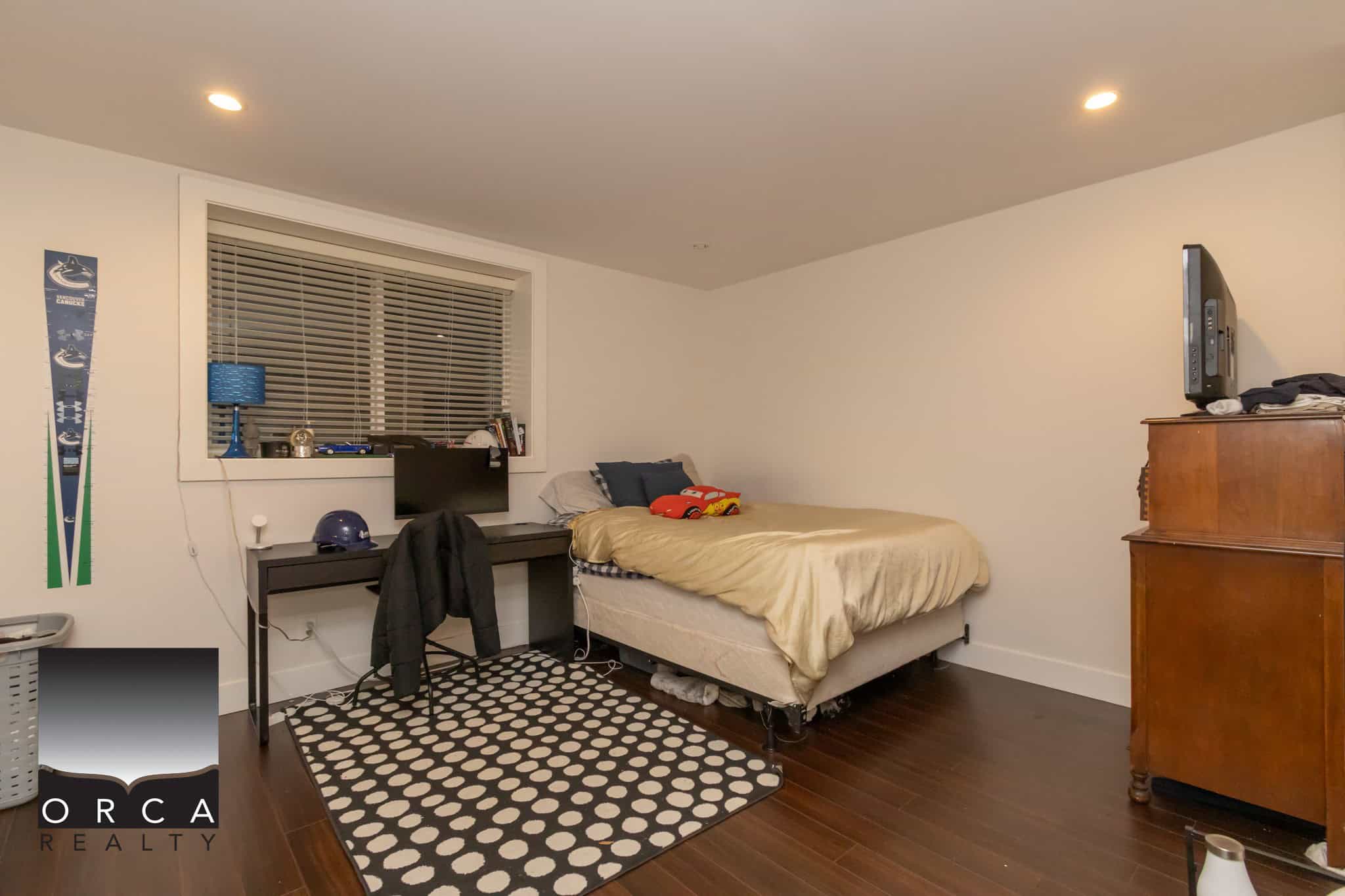 Cozy bedroom with a bed, desk, and entertainment setup, featuring dark hardwood floors and simple decor, ideal for a Vancouver Island home marketed by Orca Realty Inc.