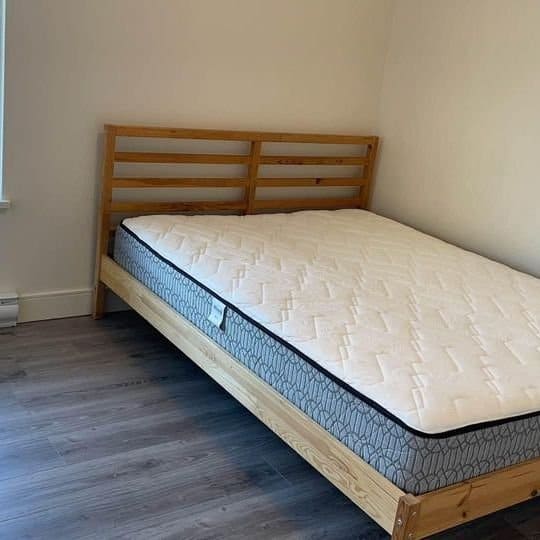 A wooden bed frame with a mattress in a simple, modern bedroom featuring laminate flooring and plain white walls.
