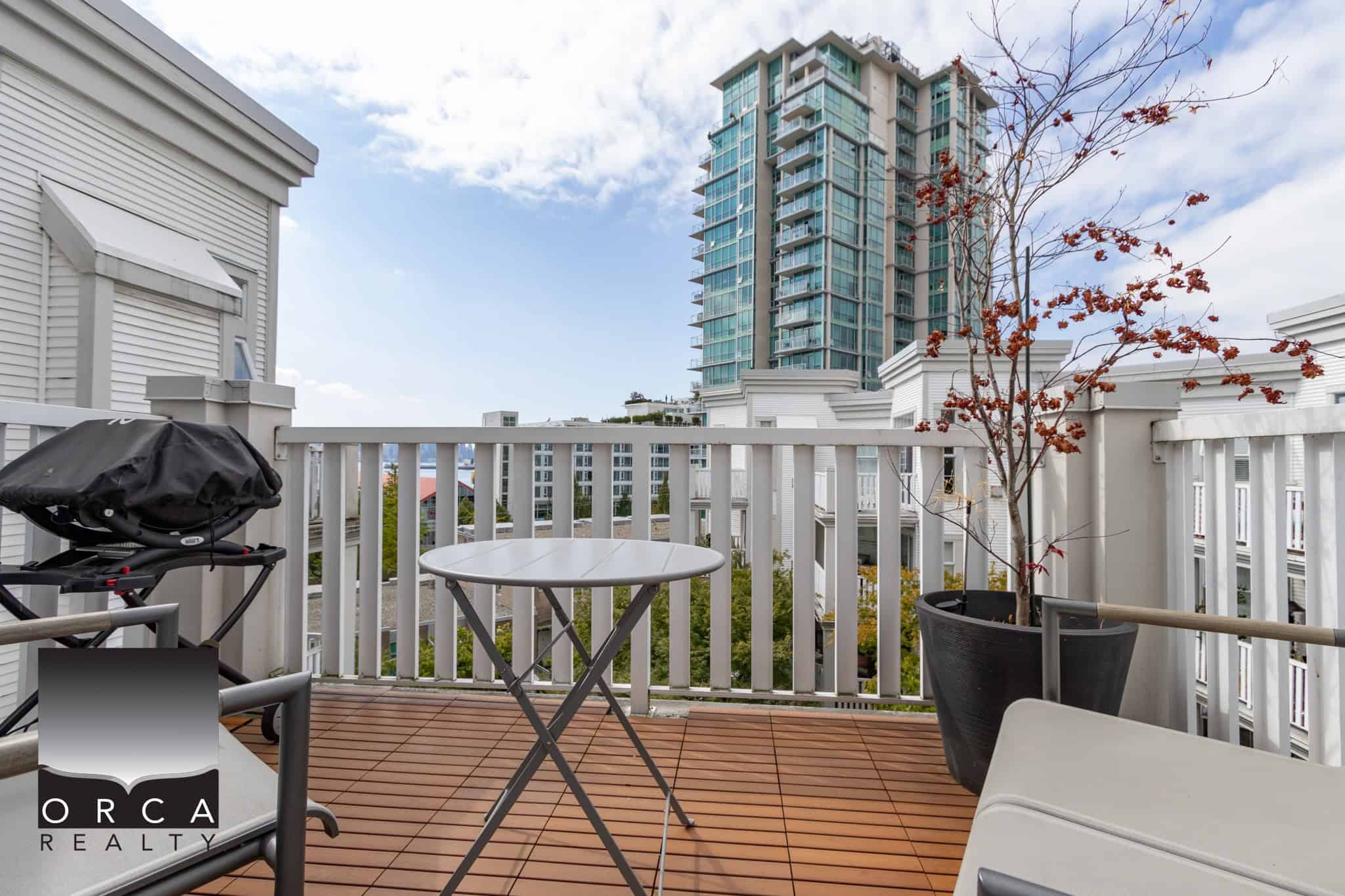 Bright urban balcony with wooden decking, potted tree, outdoor furniture, and view of modern high-rise buildings, ideal for Vancouver condo living.