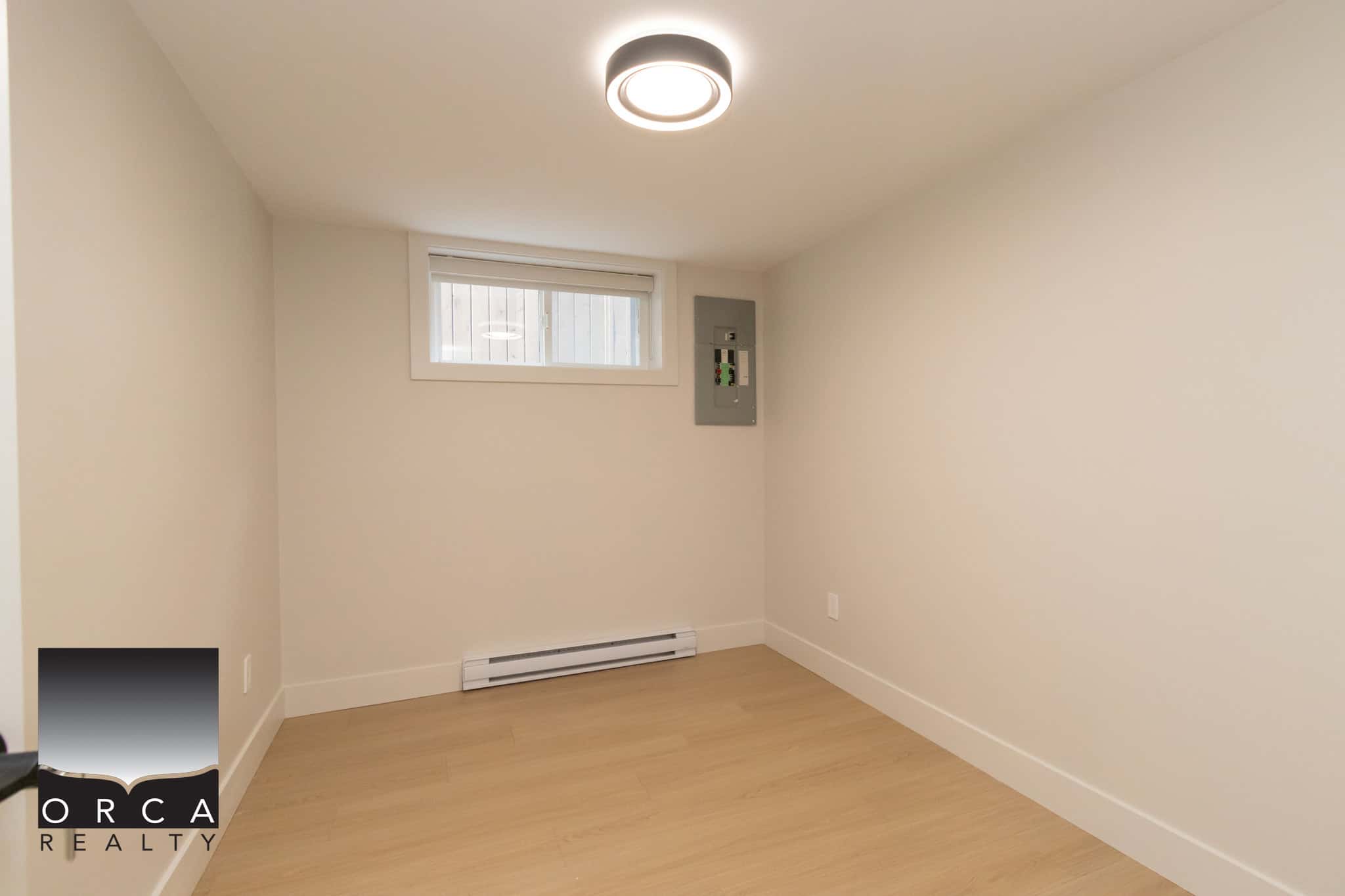 Bright empty room with a small horizontal window, neutral walls, modern ceiling light, and baseboard heater, ideal for Vancouver area homes or condos, showcasing ORCA Realty property listings.