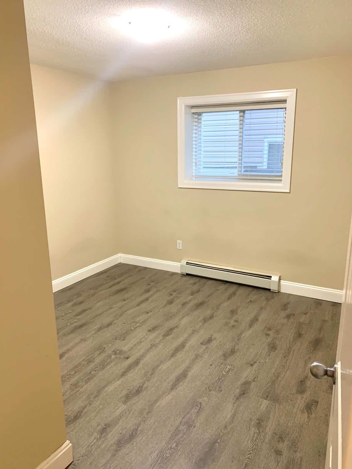 Modern empty bedroom with beige walls, white trim, and gray laminate flooring, featuring a window with blinds and a baseboard heater, ideal for Vancouver or BC real estate listings.
