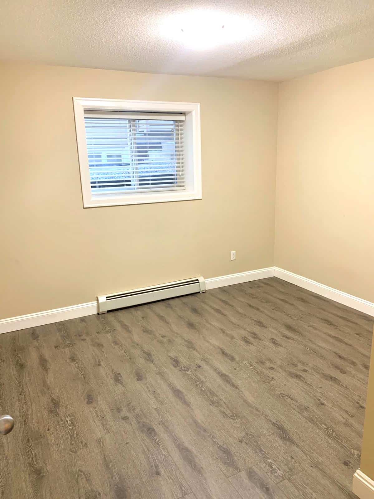 Bright empty bedroom with beige walls and vinyl plank flooring, window with white blinds, and baseboard heating, showcasing Vancouver area real estate options.
