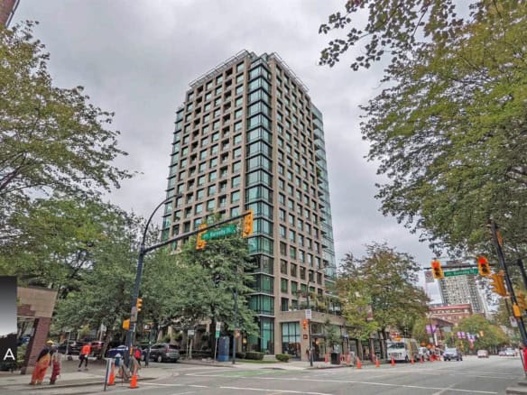 Contemporary high-rise residential building located on a busy city intersection, surrounded by lush green trees, with retail shops and traffic lights at street level, offering modern urban living options.