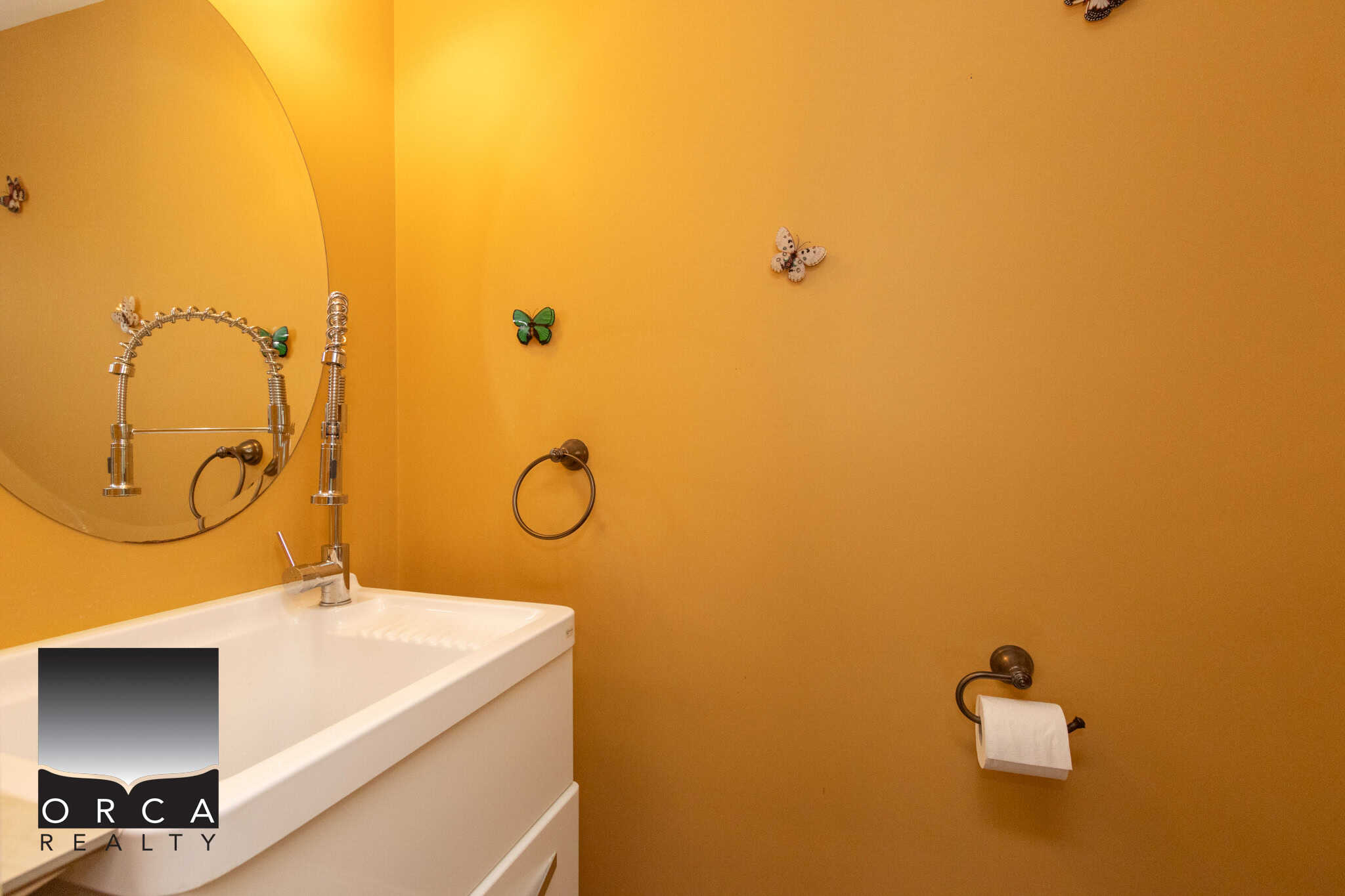 Bright yellow bathroom wall with butterfly wall decorations, a white vanity with a modern faucet, round mirror, and a towel ring, showcasing vibrant home interior design and decor.