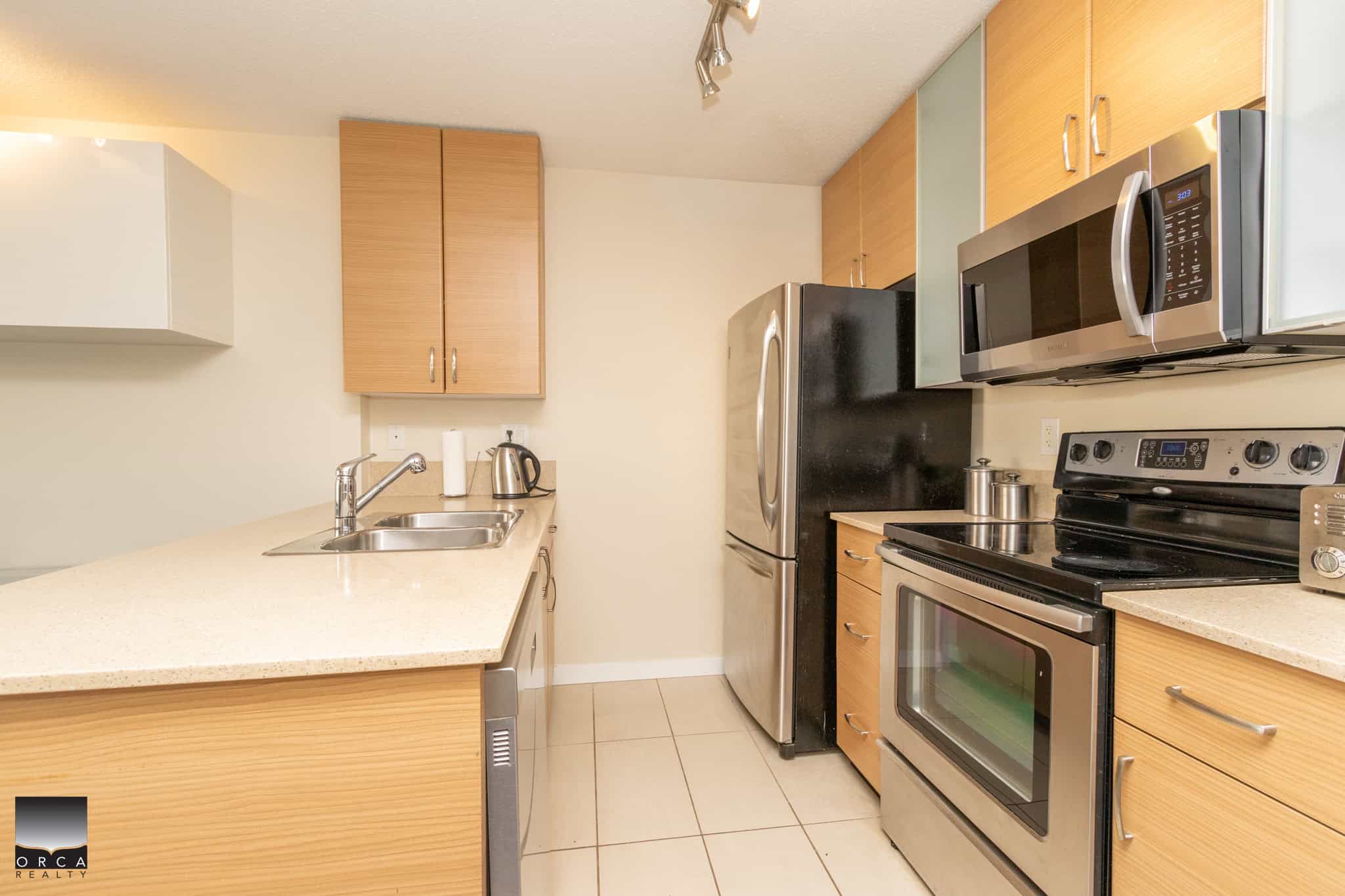 Modern fully equipped kitchen with stainless steel appliances, wooden cabinetry, beige countertops, and tiled flooring. Perfect for comfortable home living and entertaining.