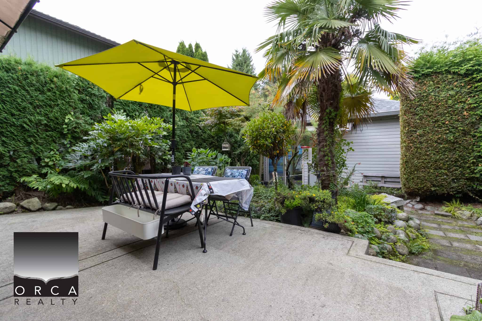 Vibrant backyard patio with outdoor dining area, lush greenery, large palm tree, and bright yellow umbrella providing shade; ideal for relaxation or entertaining guests.