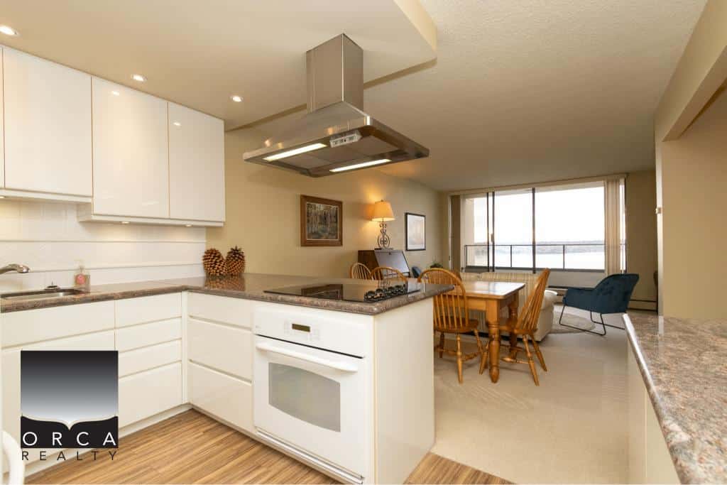 Bright modern kitchen with white cabinetry, stainless steel range hood, and open-concept living area with ocean view in a Vancouver island condo.