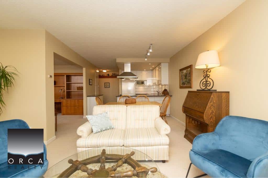 Cozy living room with beige walls, blue and cream sofas, wooden furniture, and modern kitchen in the background, showcasing Orca Realty Inc. property listings in a warm, inviting home setting.