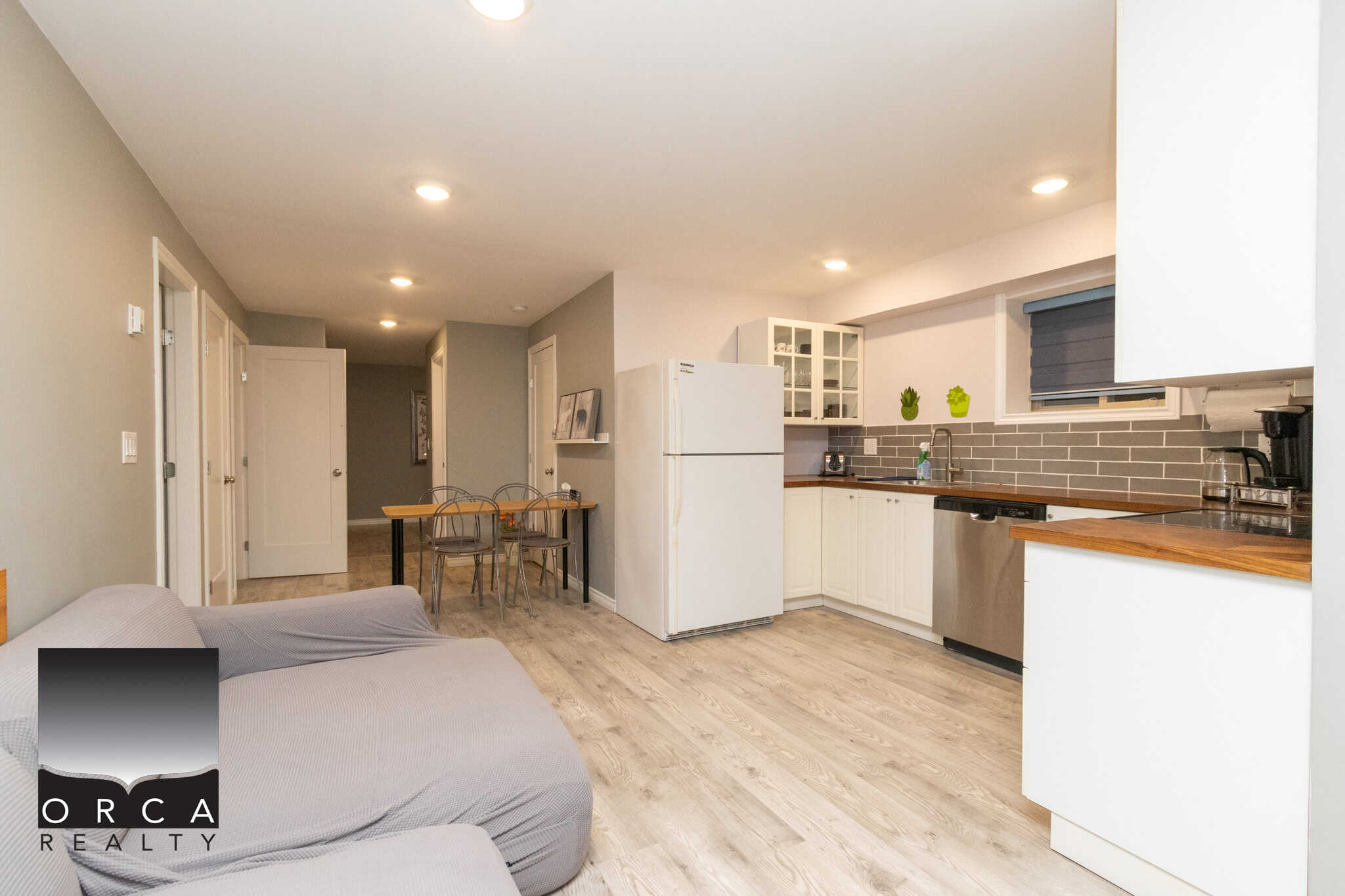 Bright modern Vancouver-style kitchen and living space in a contemporary condo by Orca Realty Inc., featuring white cabinetry, stainless steel appliances, and light hardwood flooring.