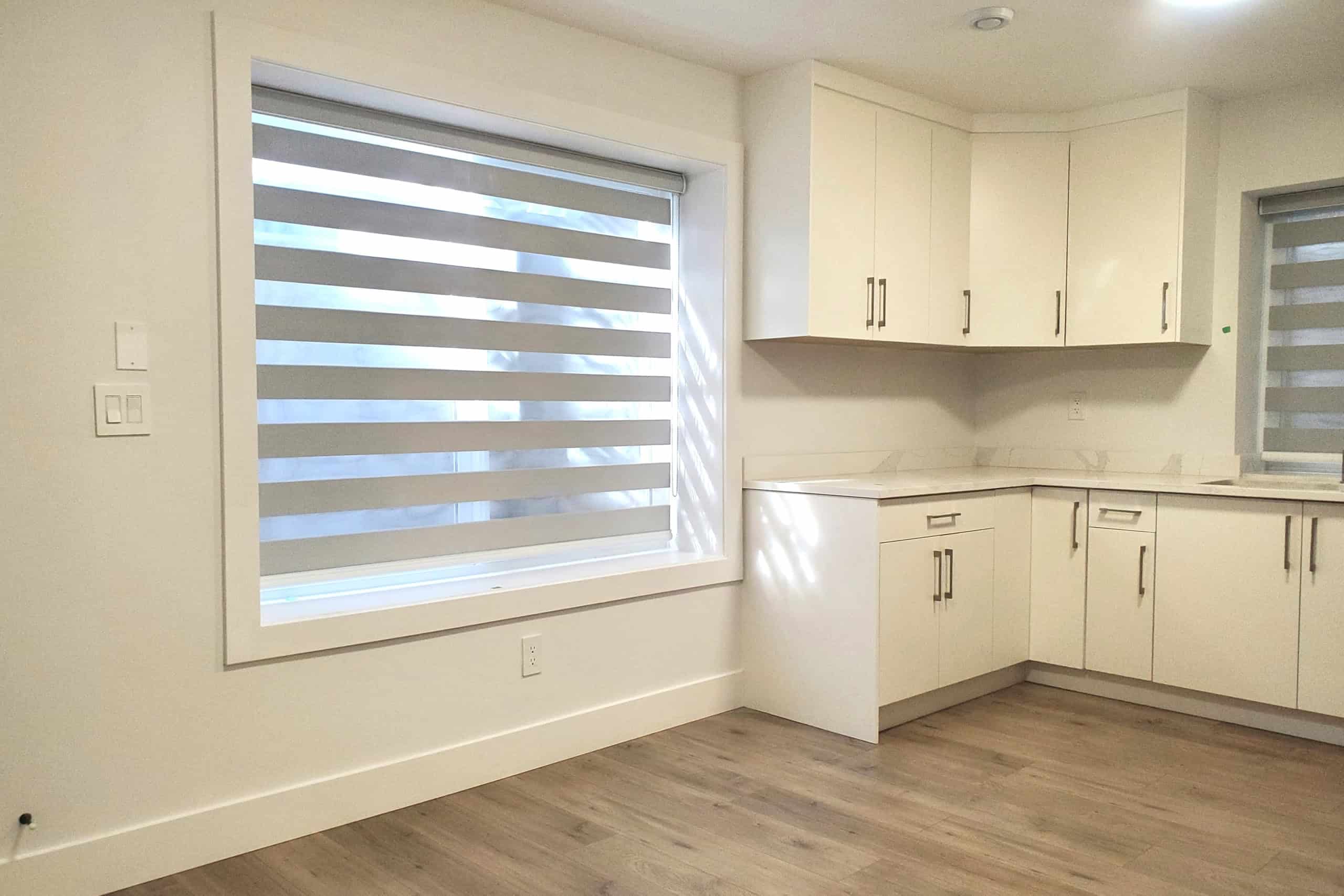 Bright modern kitchen with white cabinets, marble countertops, and large windows with zebra blinds, showcasing contemporary home design and natural light.