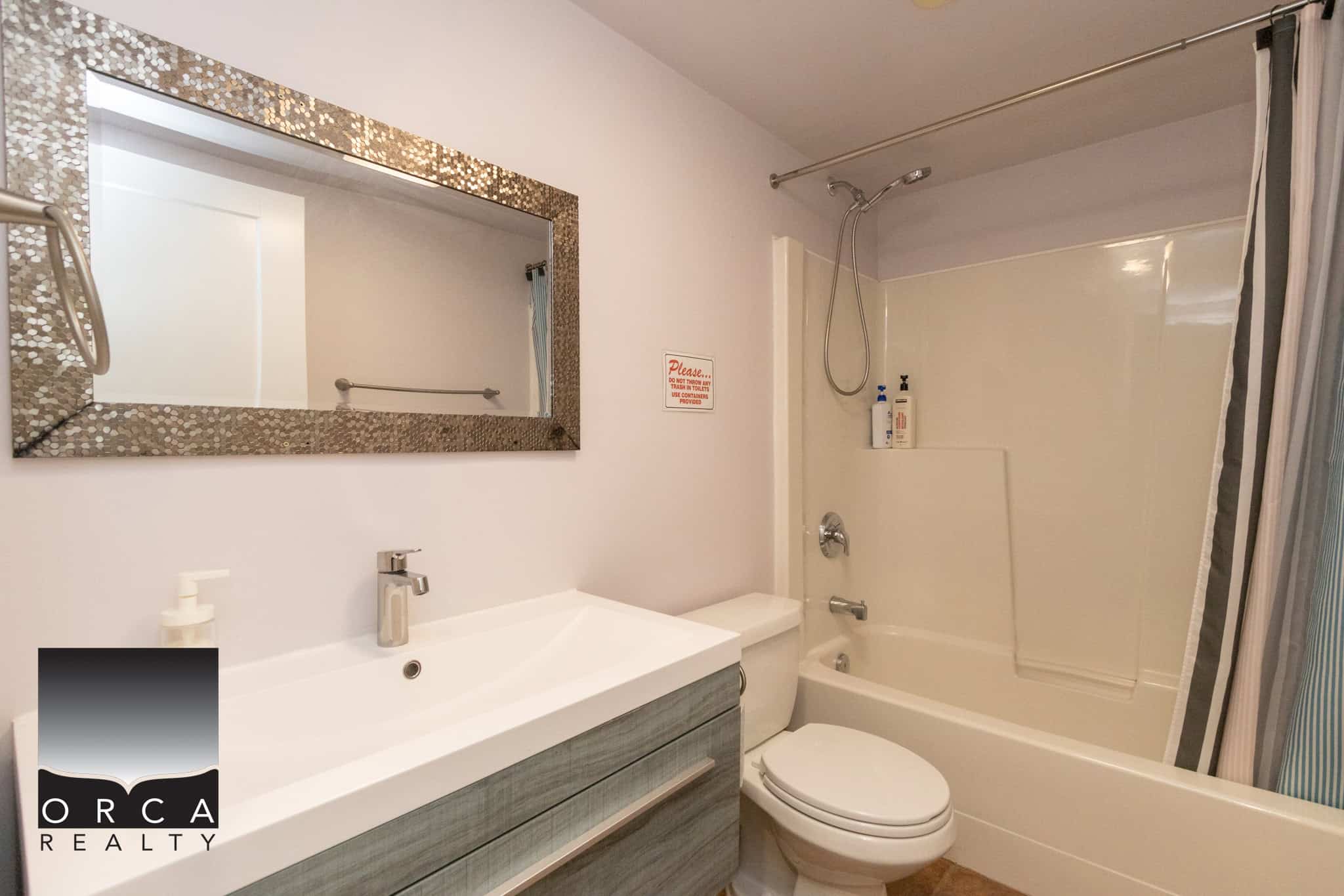 Clean modern bathroom with a shower/tub combo, white fixtures, and a large decorative mirror, ideal for Vancouver island homes and condos marketed by Orca Realty Inc.