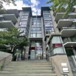 Modern Vancouver condominium building with glass facade and outdoor balconies, surrounded by lush trees, under a bright blue sky with scattered clouds.