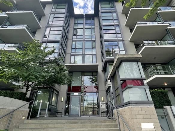Modern Vancouver condominium building with glass facade and outdoor balconies, surrounded by lush trees, under a bright blue sky with scattered clouds.