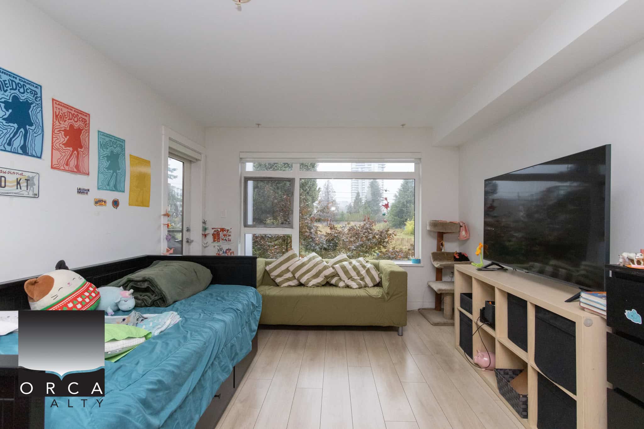 Cozy living room with large windows showcasing greenery, featuring a black bed frame, green sofa, TV on a wooden stand, and colorful wall art, perfect for Vancouver real estate.