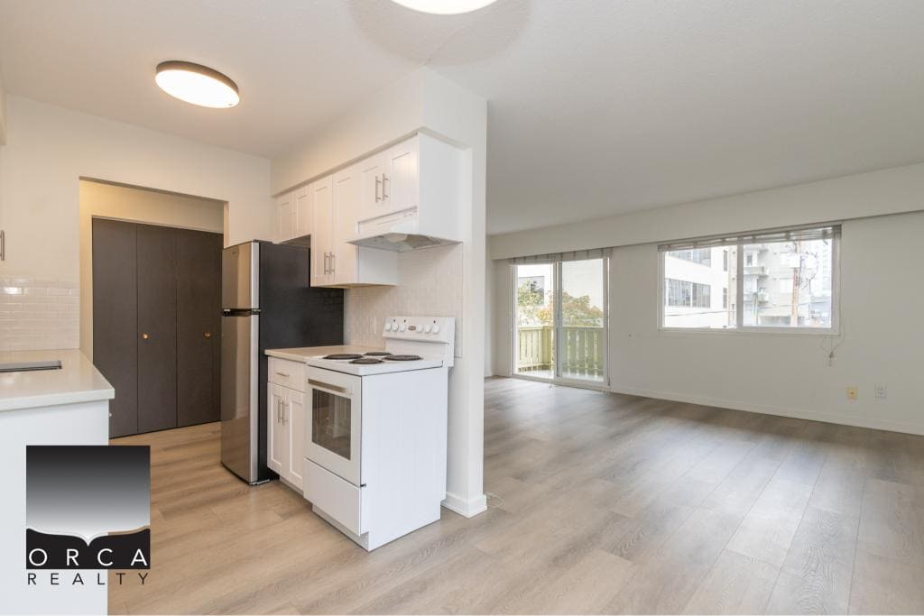 Modern open-concept living room and kitchen in Vancouver apartment, featuring large windows, bright white cabinetry, and stainless steel appliances, ideal for comfortable downtown living.